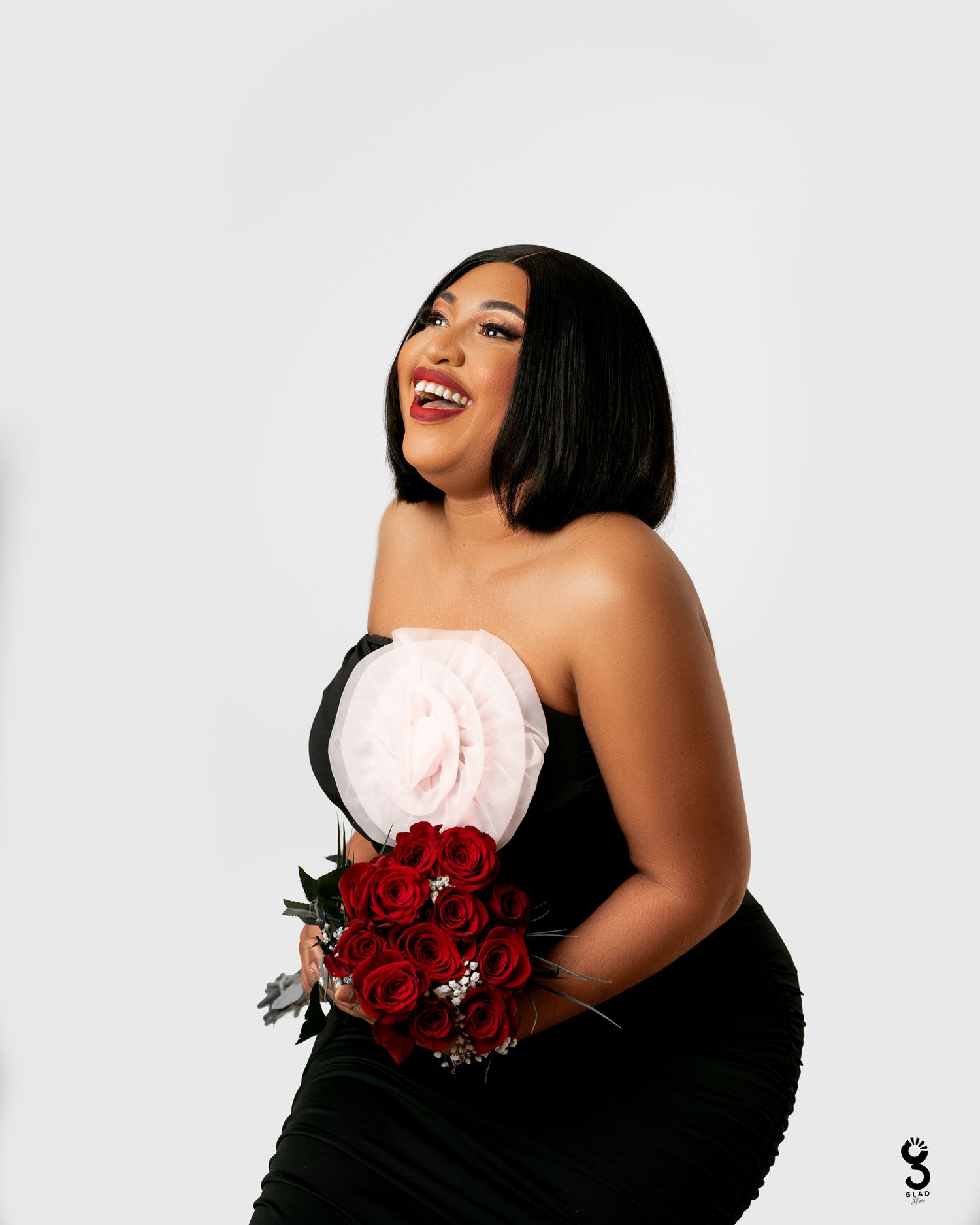 A person in a black strapless dress poses with red roses and a large pink floral decoration against a white backdrop. birthday photos