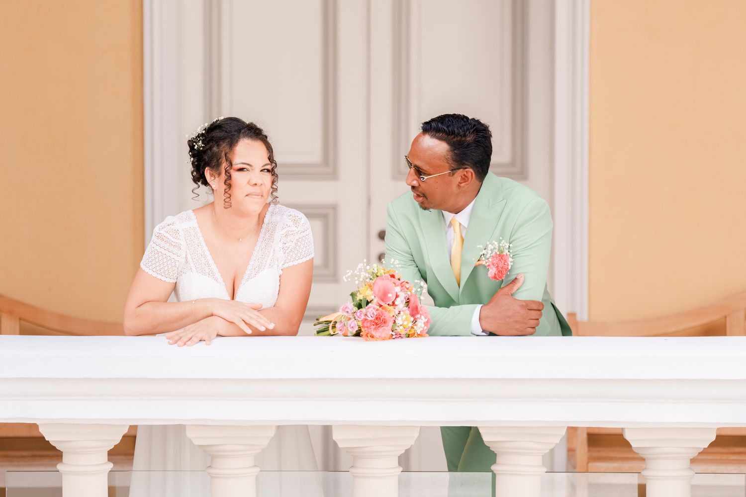 A couple sits together at an elegant white balcony railing with pink flowers and romantic lighting during a formal occasion.
