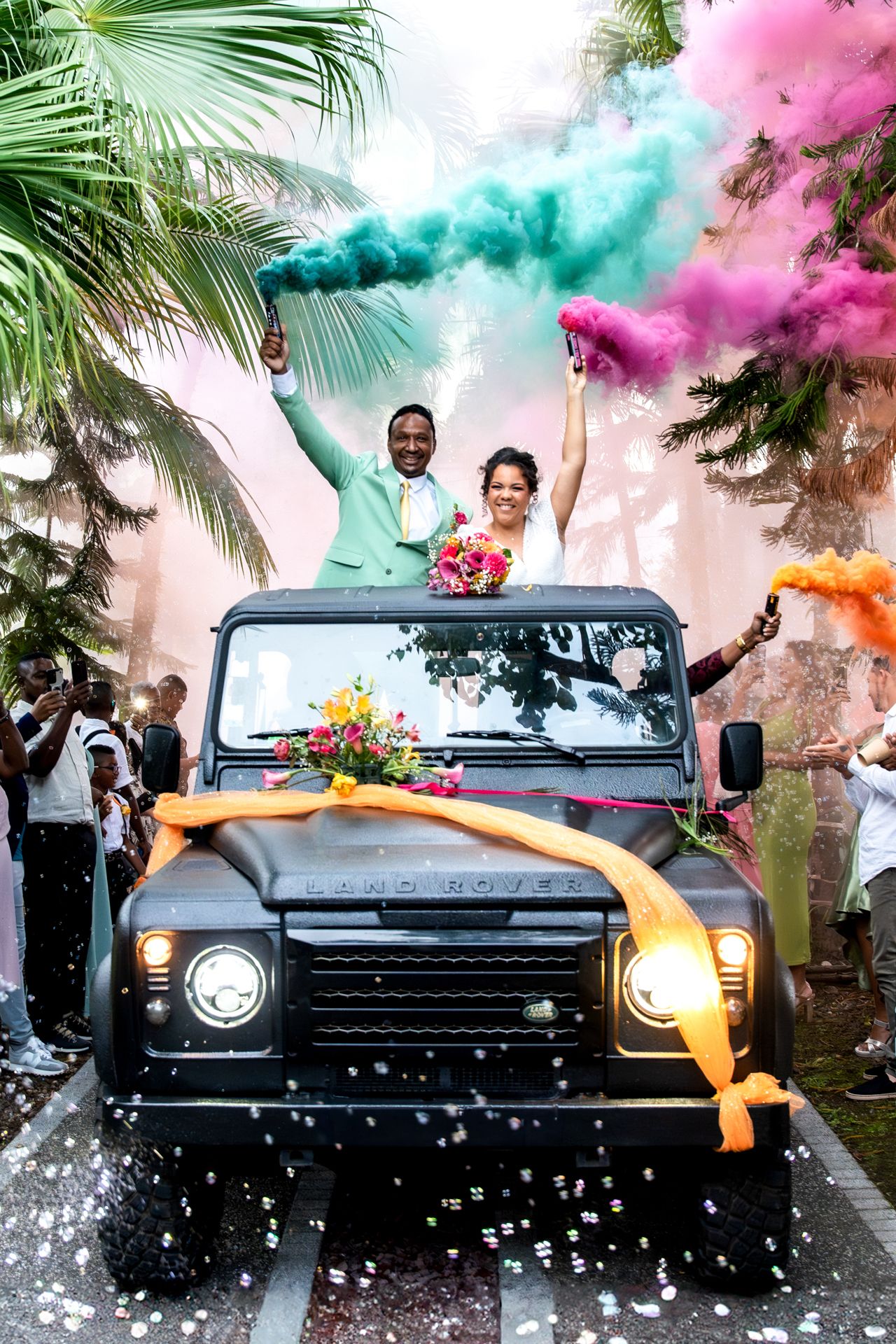 A couple celebrates in a decorated black Defender SUV with colorful smoke bombs and palm trees in the background.