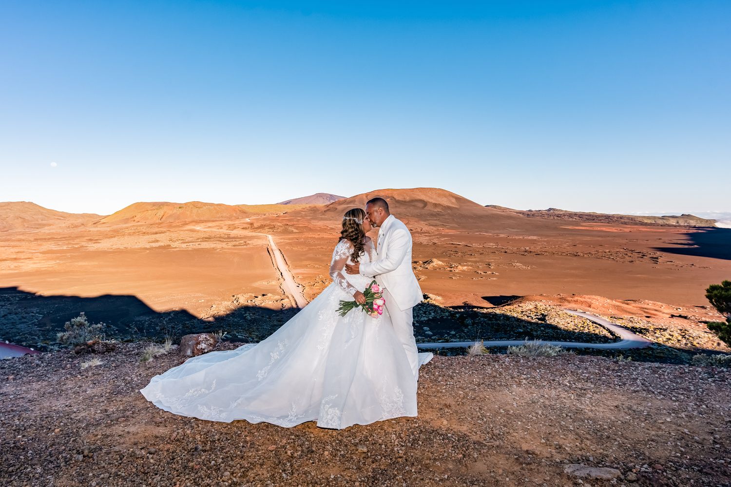 A wedding couple stands on rugged desert terrain against orange mountains and bright blue sky.