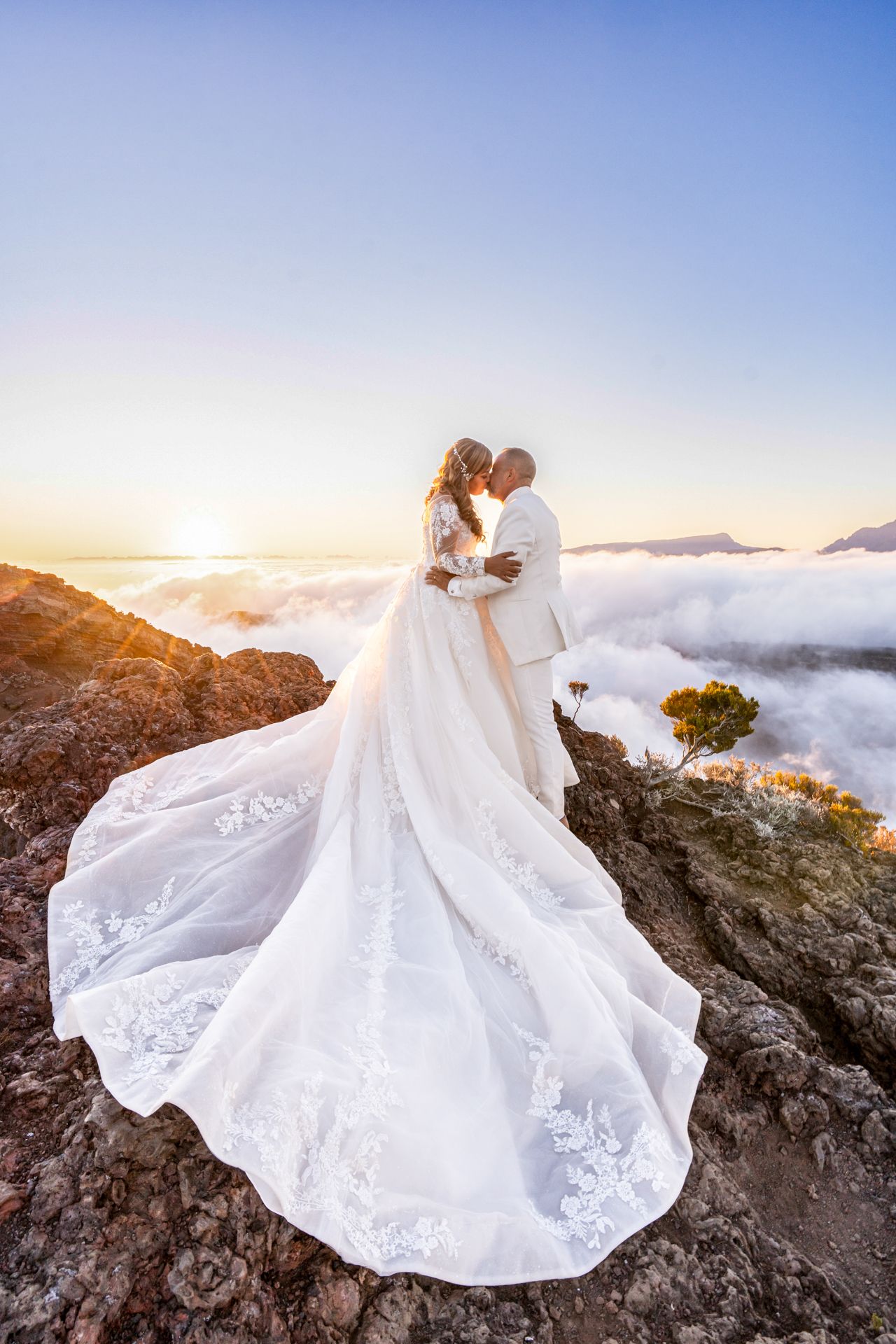 A wedding couple embraces on a mountain peak above the clouds during sunset, with a flowing white lace gown trailing behind.