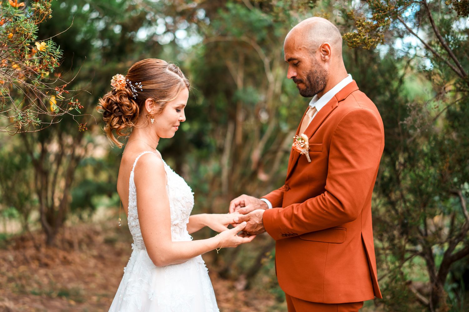 A bride wearing a white dress and their partner in a burnt orange suit exchange rings during an outdoor wedding ceremony.