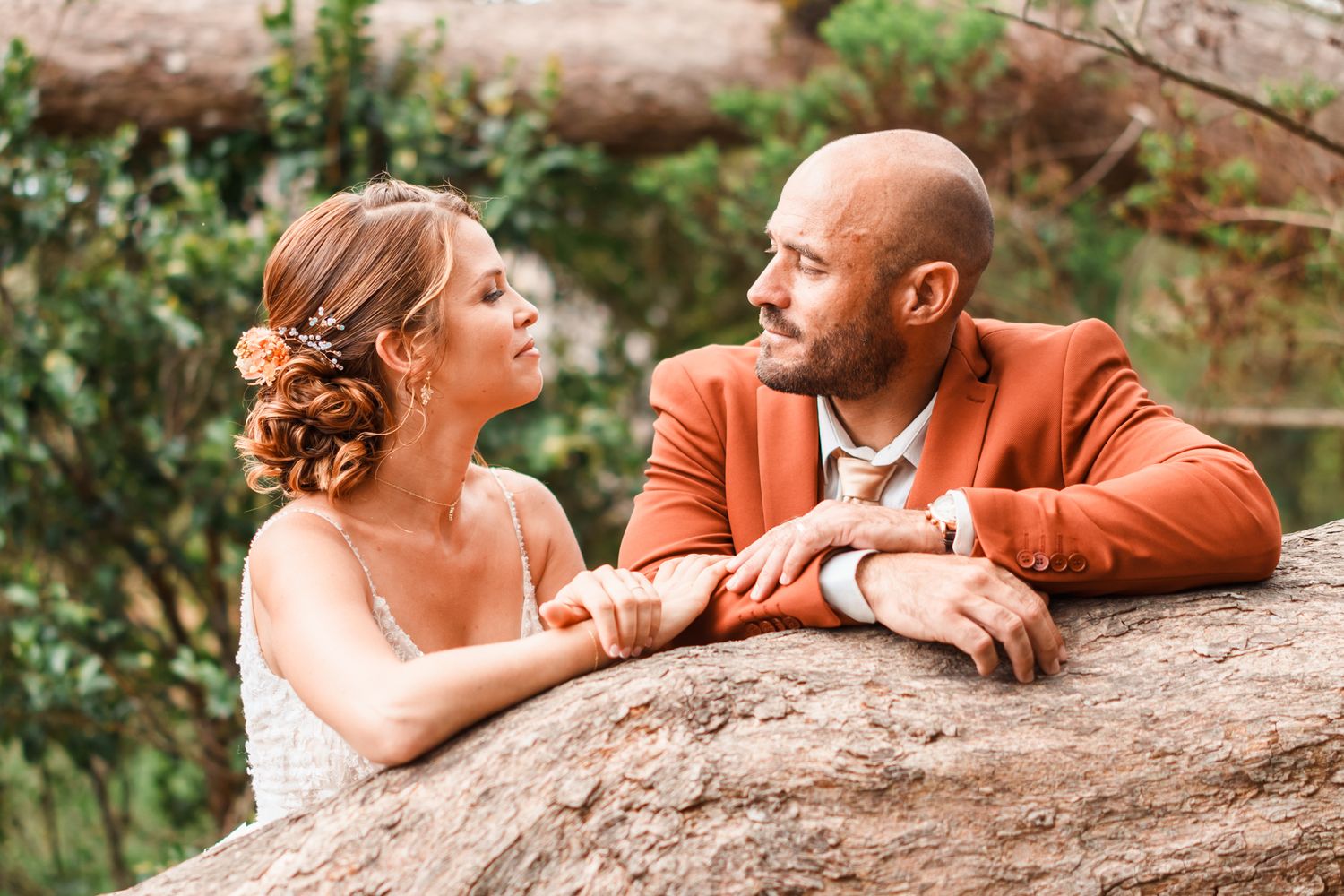 A couple in wedding attire gazes at each other while leaning on a large rock in an outdoor garden setting.