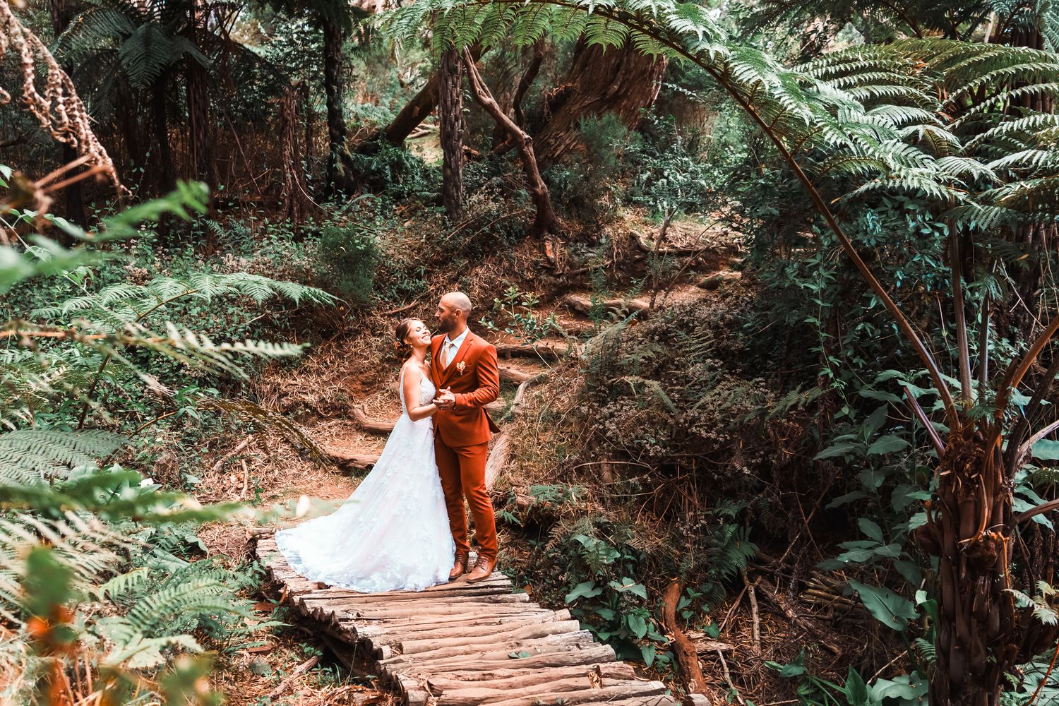 A couple in wedding attire stands on a wooden footbridge surrounded by lush ferns and forest greenery.