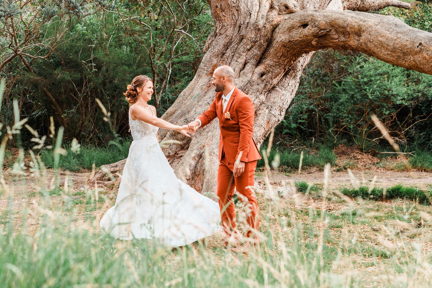 A couple in white lace gown and bright burnt orange suit dancing outdoors near a large tree trunk.
