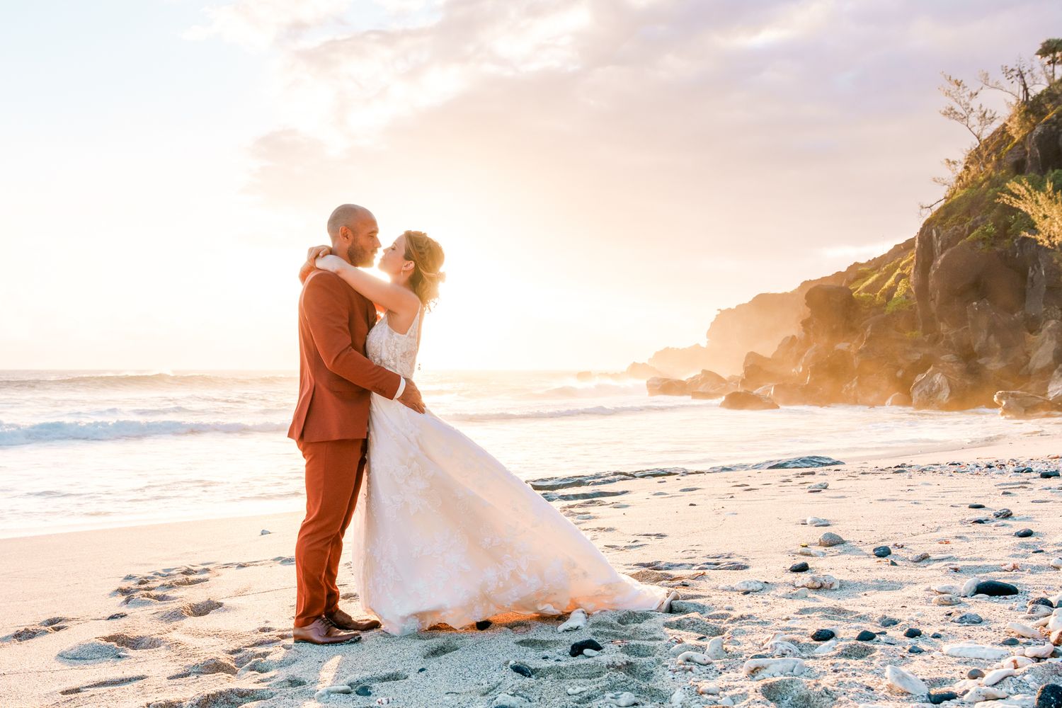 A couple in wedding attire embraces on a scenic beach at sunset with cliffs and crashing waves in the background.