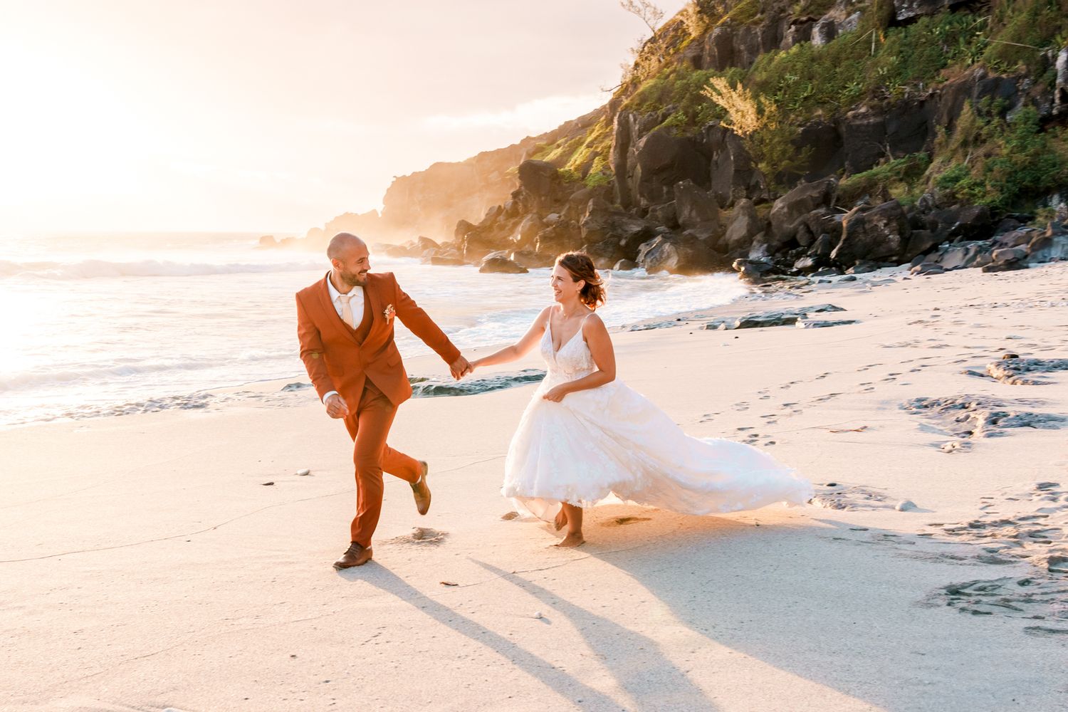 A couple in wedding attire running and holding hands on a scenic beach at sunset with cliffs in the background.