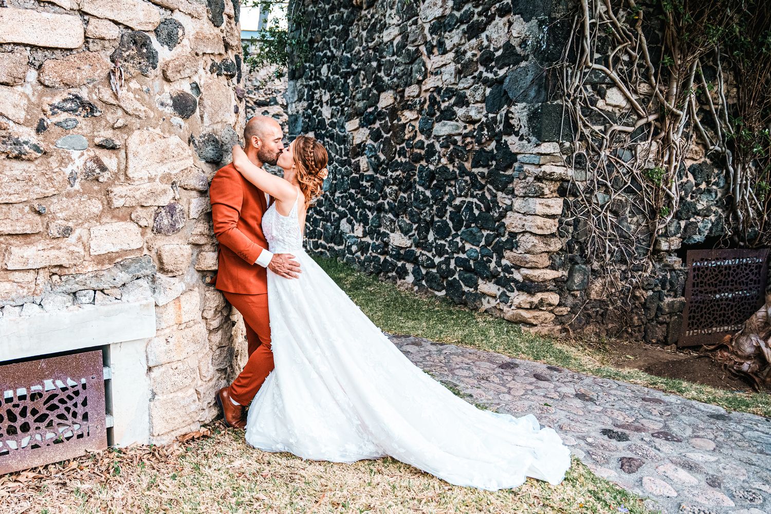 A white wedding gown and orange suit contrast against a rustic stone wall on a natural path.