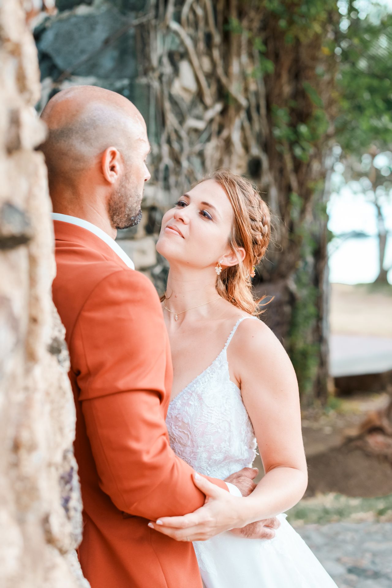 Romantic wedding moment captured between couple in white bridal gown and rust-colored suit embracing by stone wall outdoors.