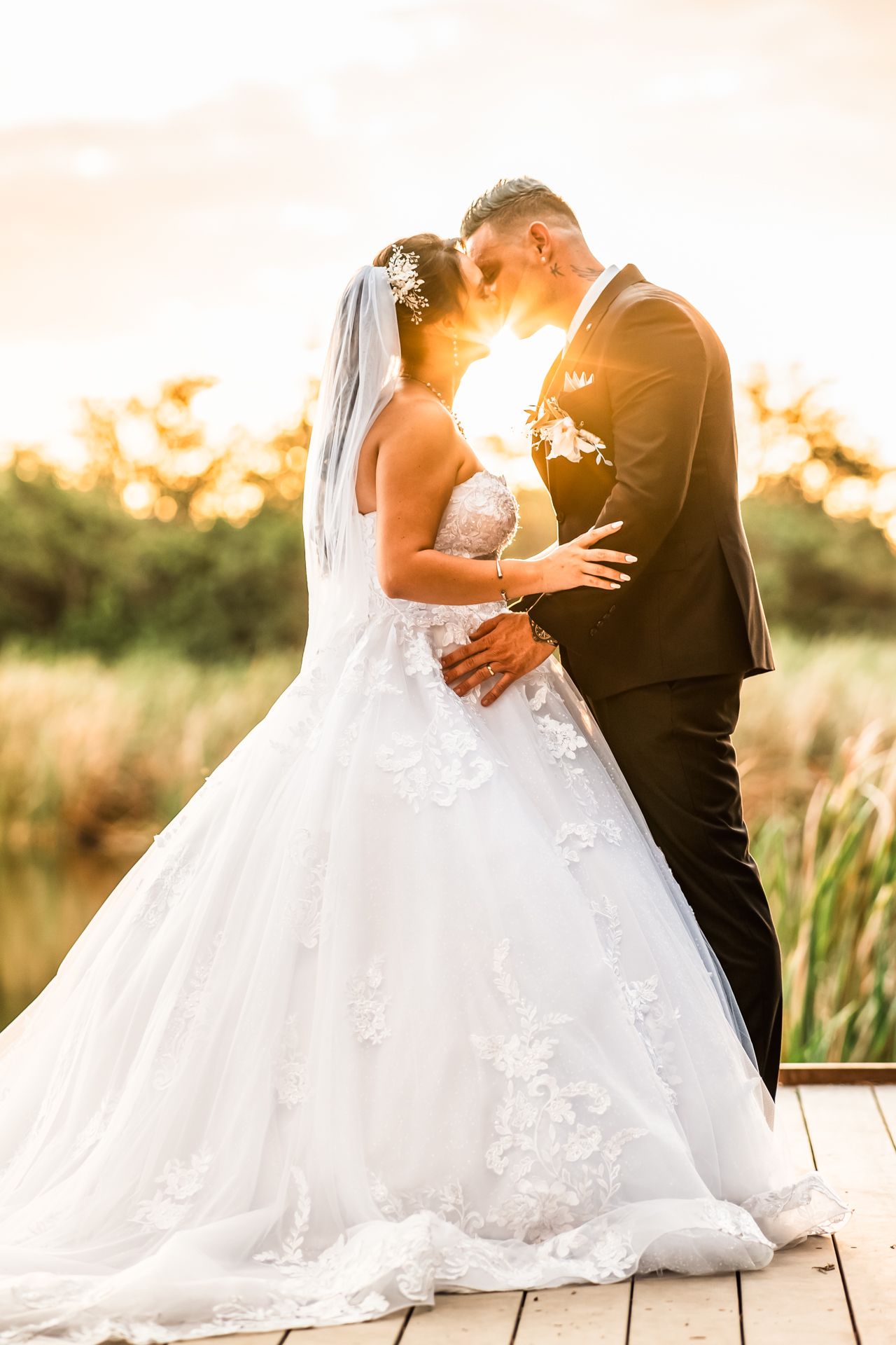 Bride and groom share intimate sunset moment on wooden deck with lush greenery in background.