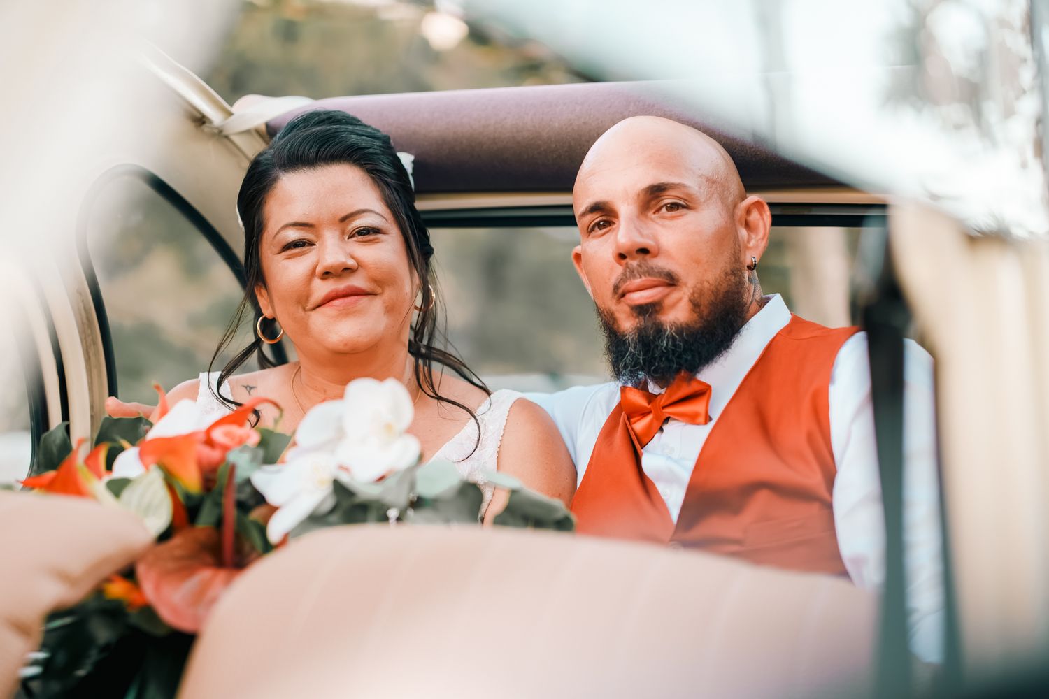 A bride and groom share an intimate moment in a wedding car, with the bride holding a white floral bouquet.