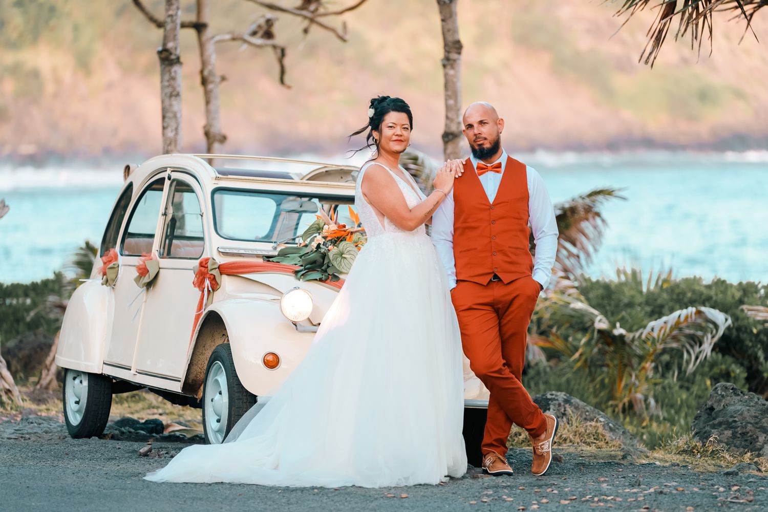 A couple poses by a vintage cream-colored Citroen 2CV car at their waterfront wedding celebration.