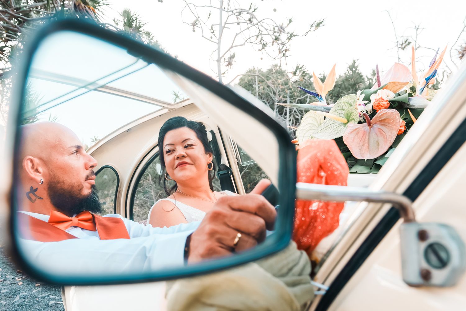 Side mirror reflection shows couple enjoying a road trip in their car with coral colored flowers visible outside.