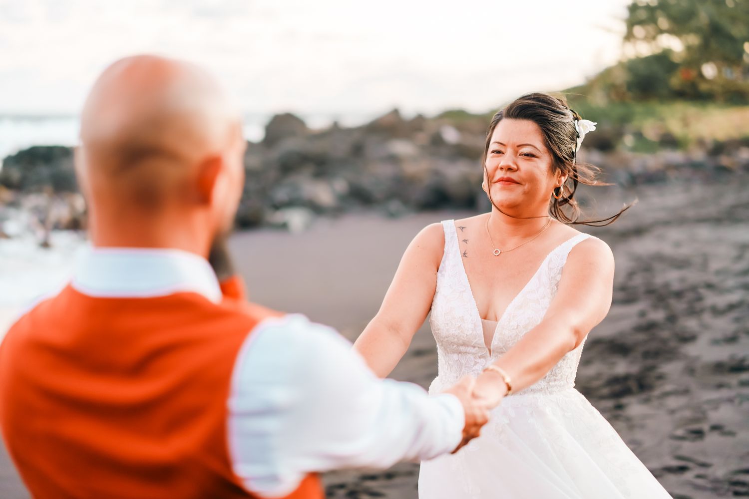 Joyful couple in wedding attire hold hands while dancing together on a black sand beach at sunset.