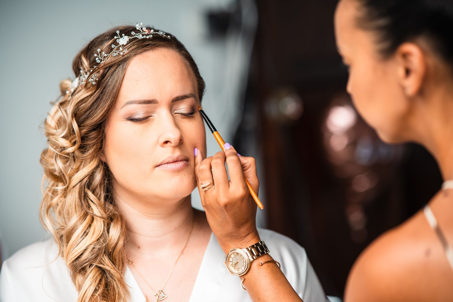 A bride getting her makeup professionally applied while wearing a white robe and crystal hairpiece.
