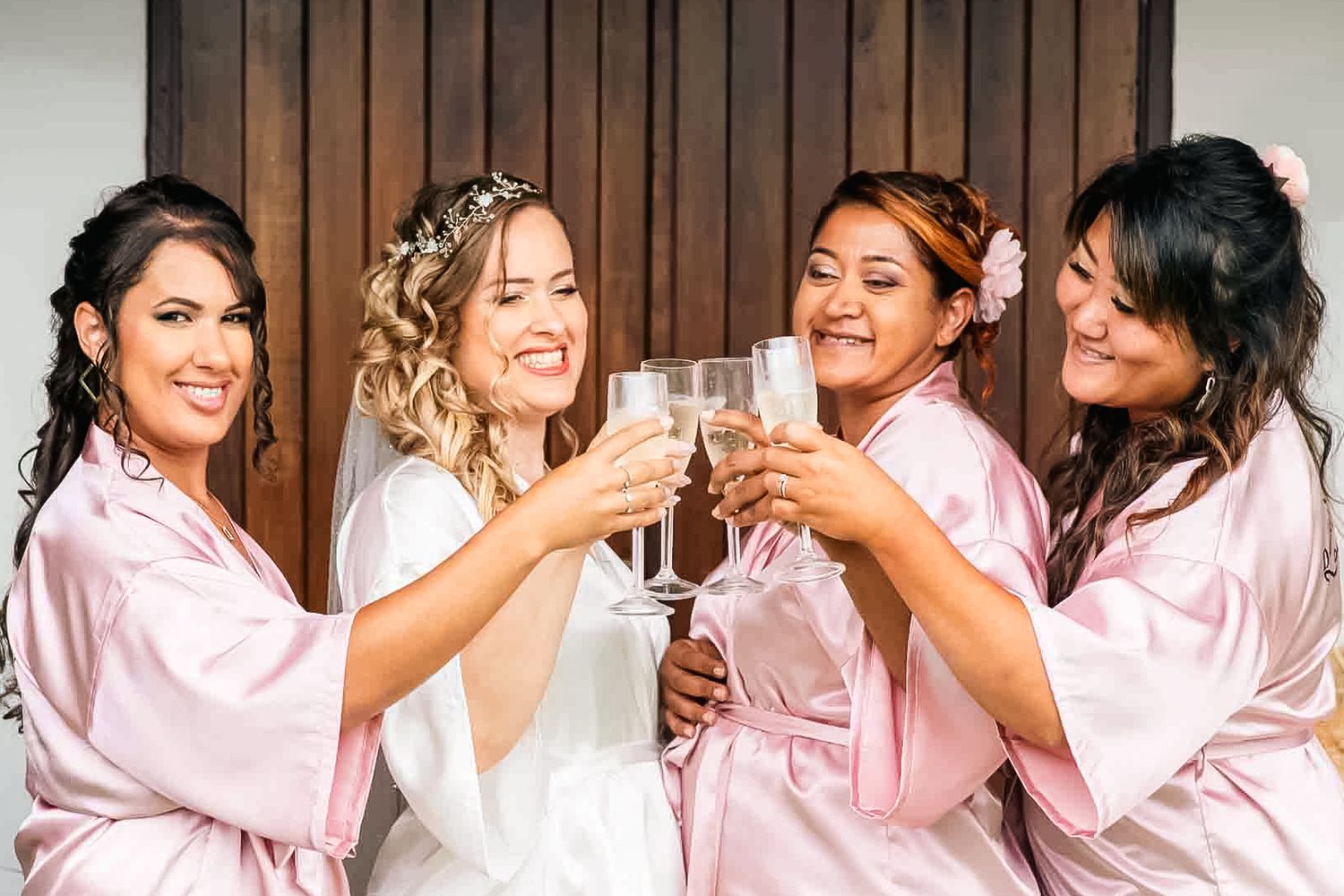Group celebrates in matching pink silk robes raising champagne glasses in a cheers moment at pre-wedding gathering.