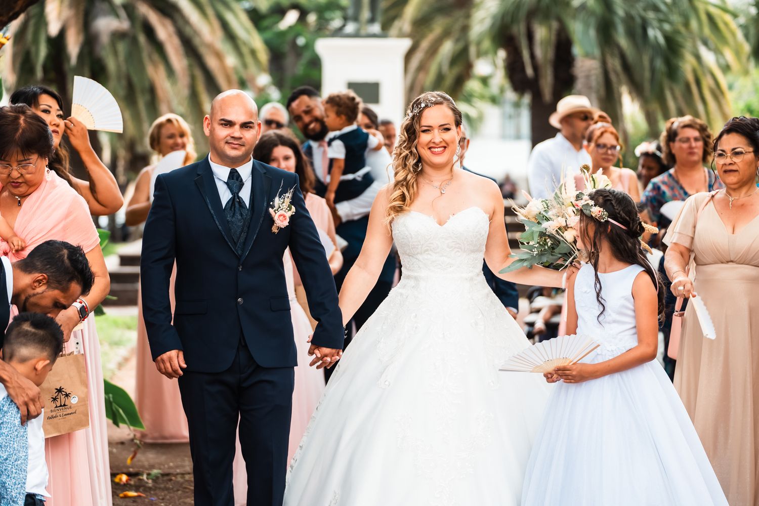 A wedding ceremony in an outdoor garden setting with palm trees and elegantly dressed guests in attendance.