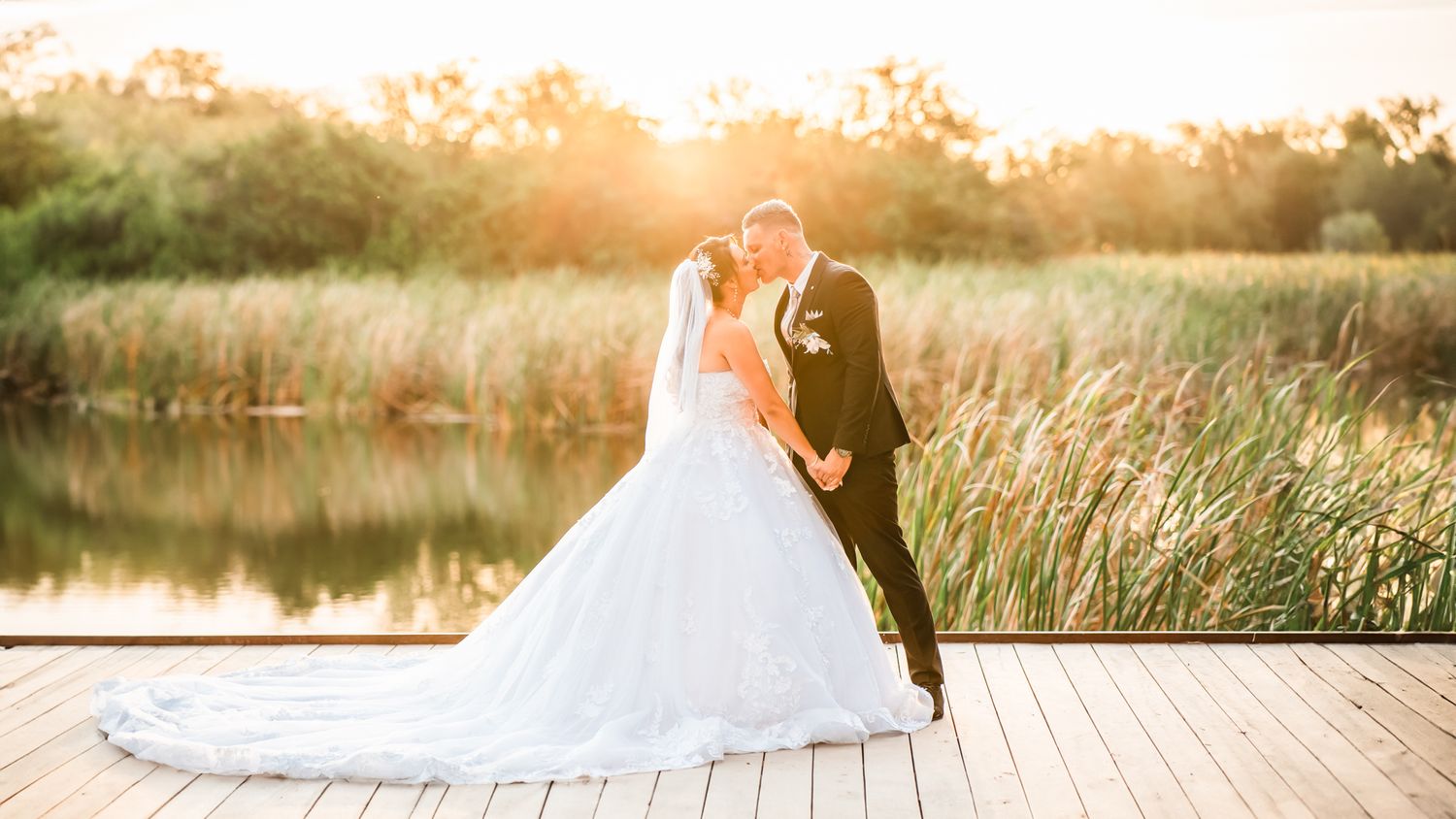 Newlywed couple embraces on wooden dock at sunset with lake and golden grass in background.