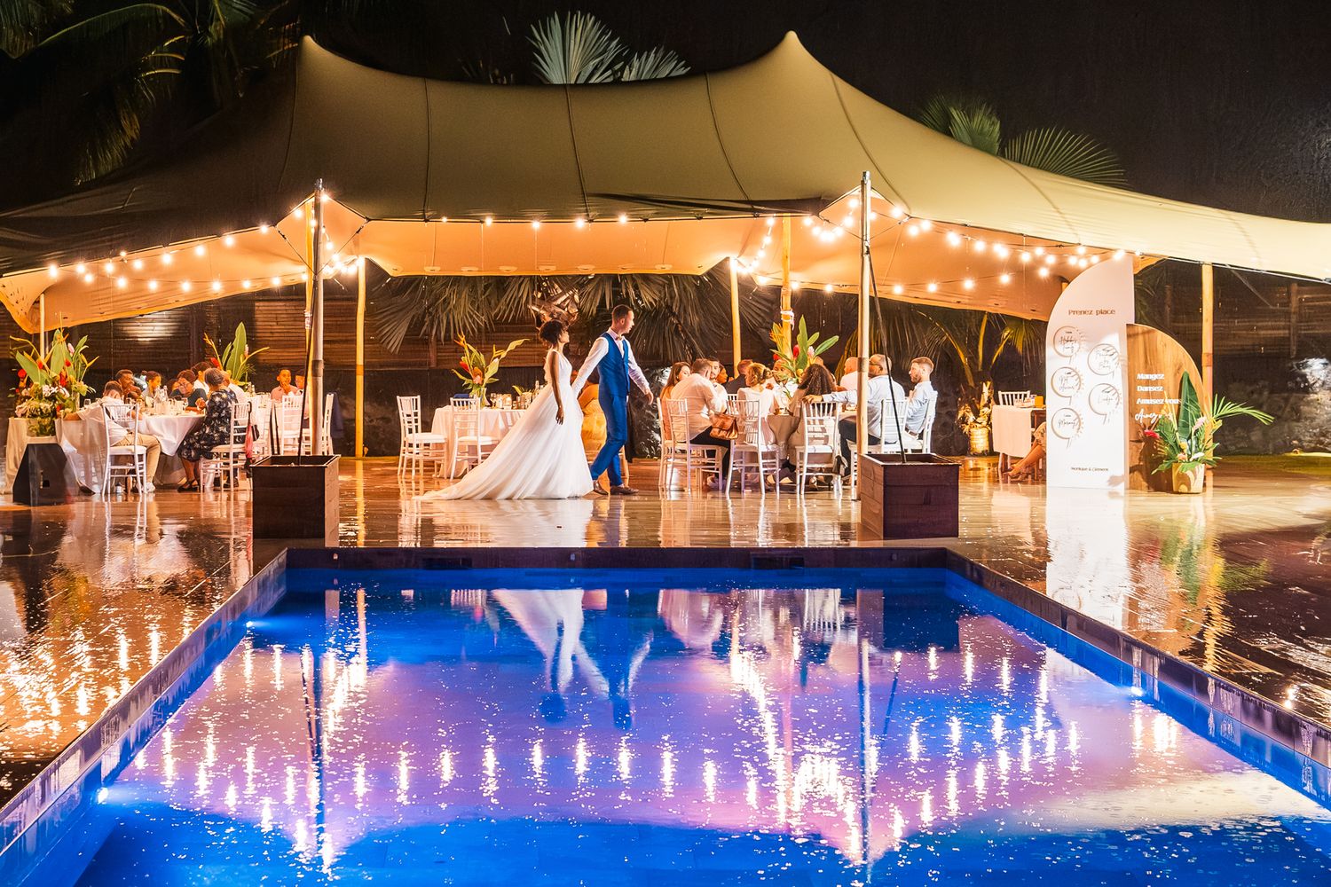 Elegant wedding reception under a lit tent with people dancing beside a glowing blue swimming pool at night.