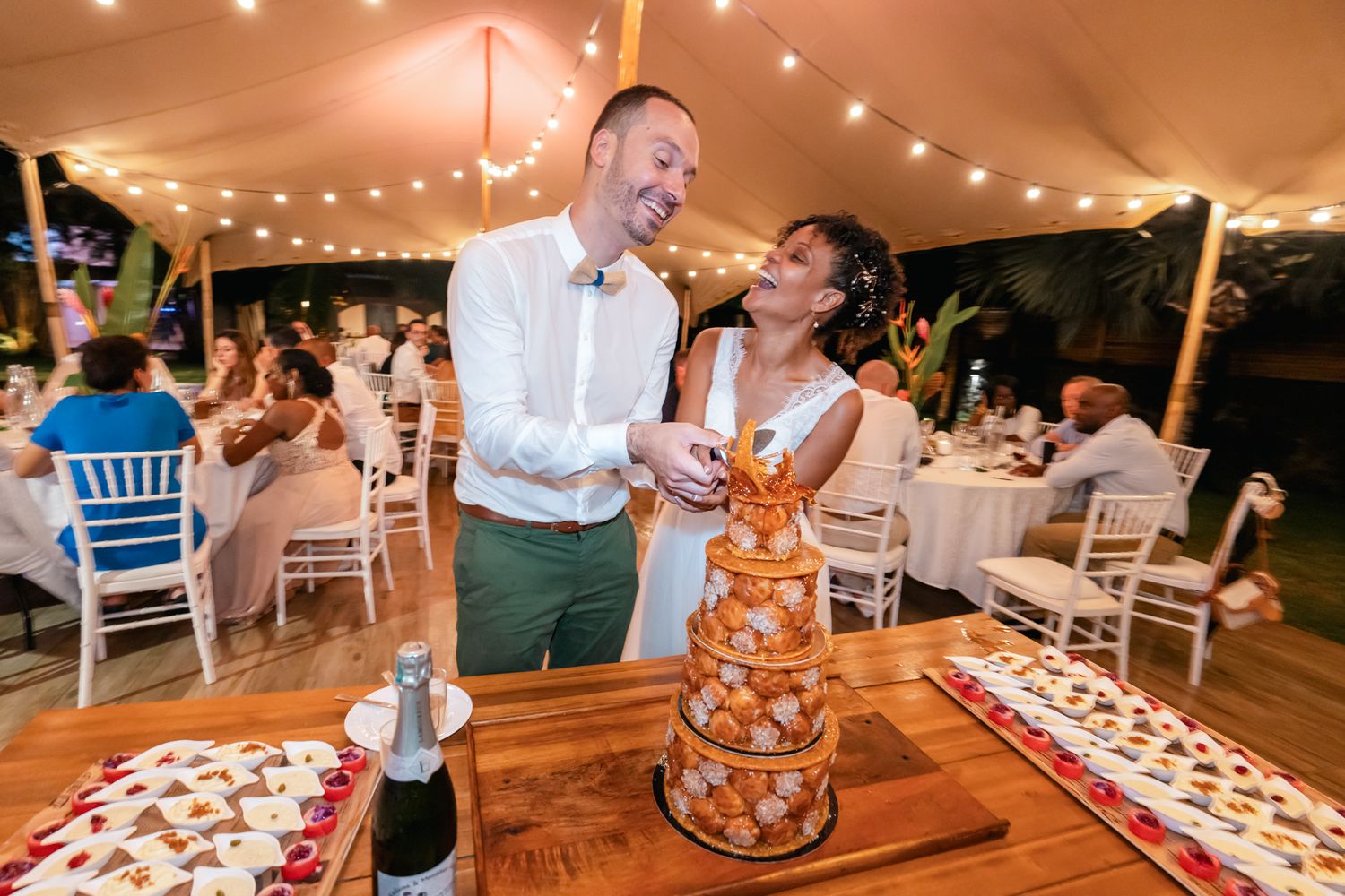 A wedding couple shares a joyful moment while cutting their dessert under a tent with twinkling lights and white chairs.