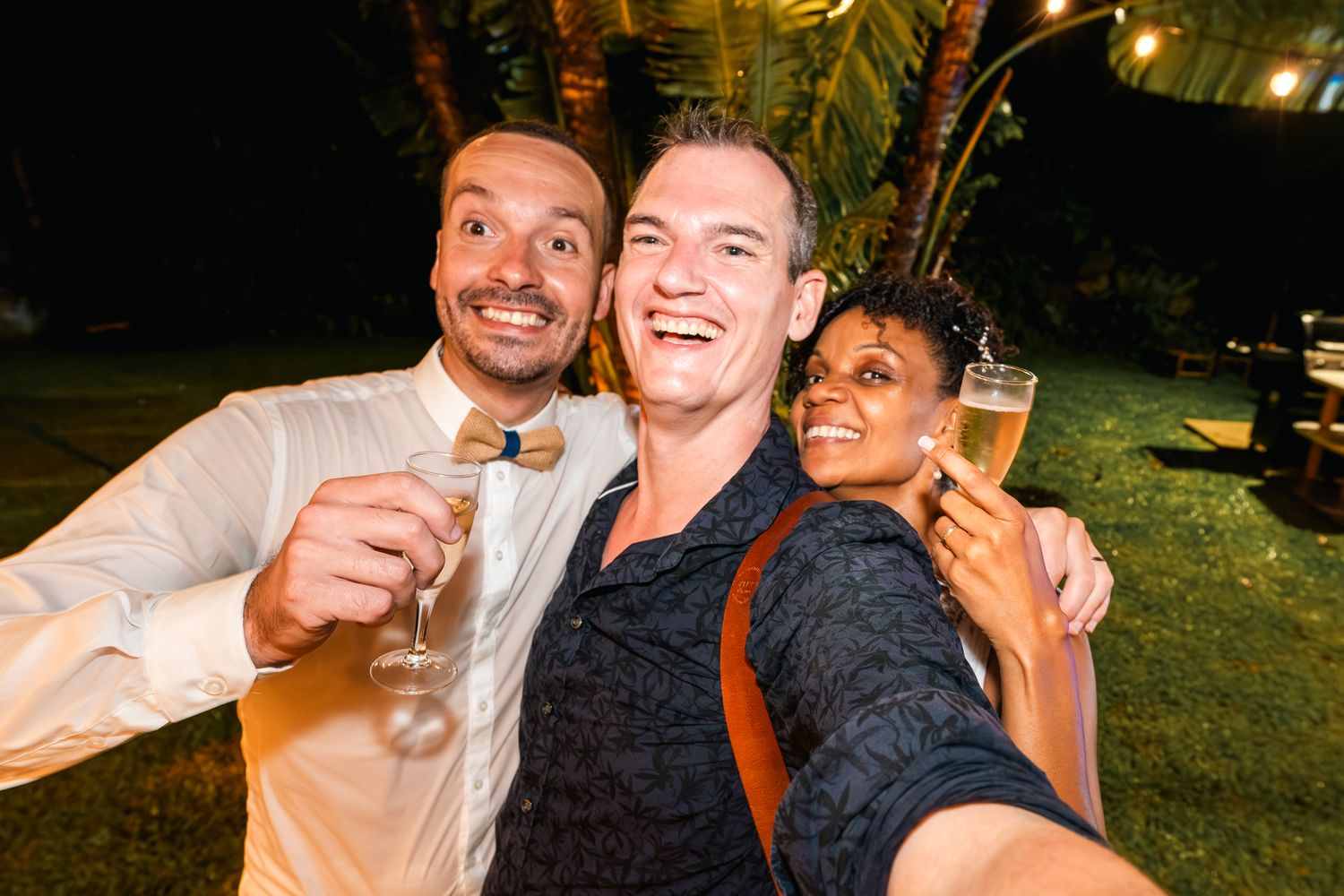 Three friends share drinks and smiles while taking a selfie together at an outdoor evening party.