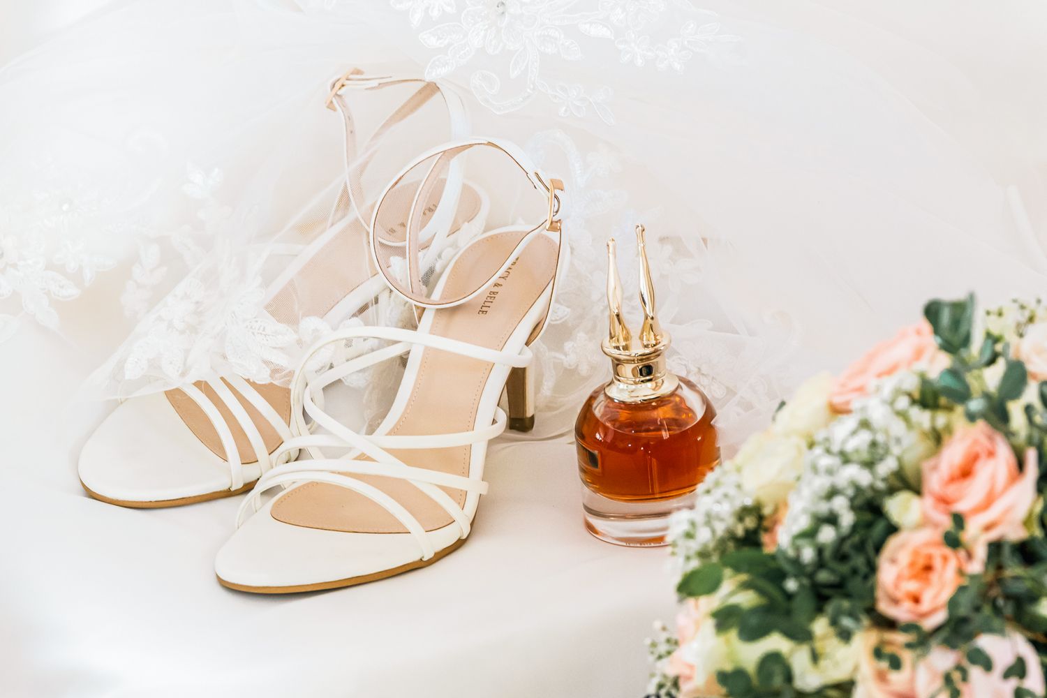 Elegant white strappy wedding sandals next to a perfume bottle and pink floral bouquet arrangement on white surface.