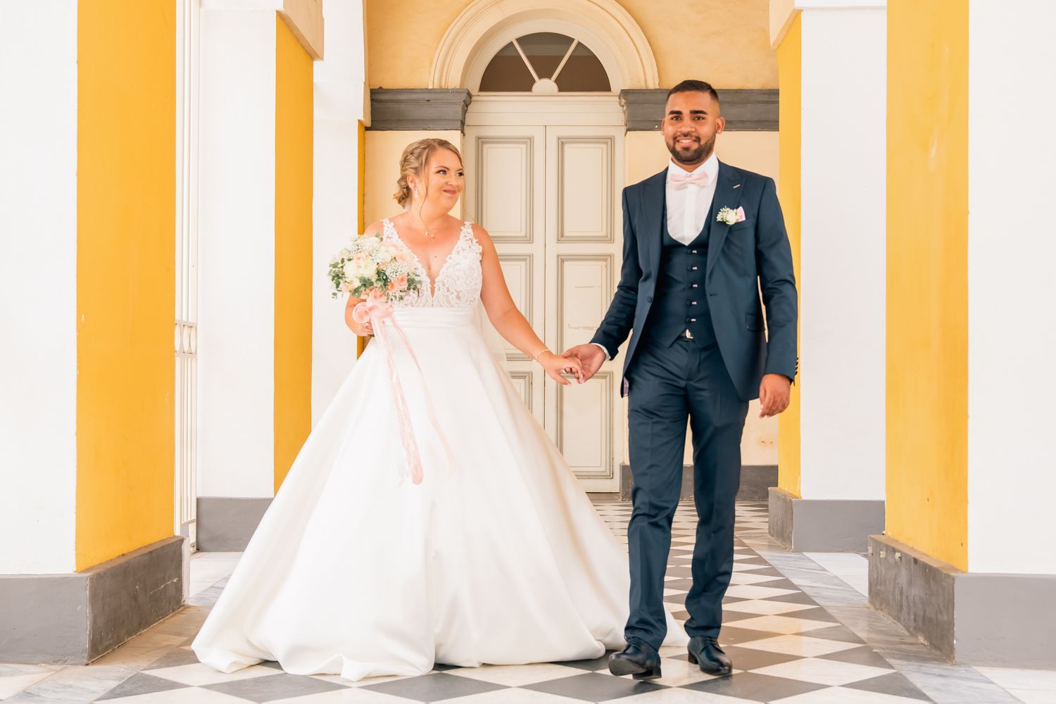 A couple stands together on their wedding day in front of a white door with yellow columns.