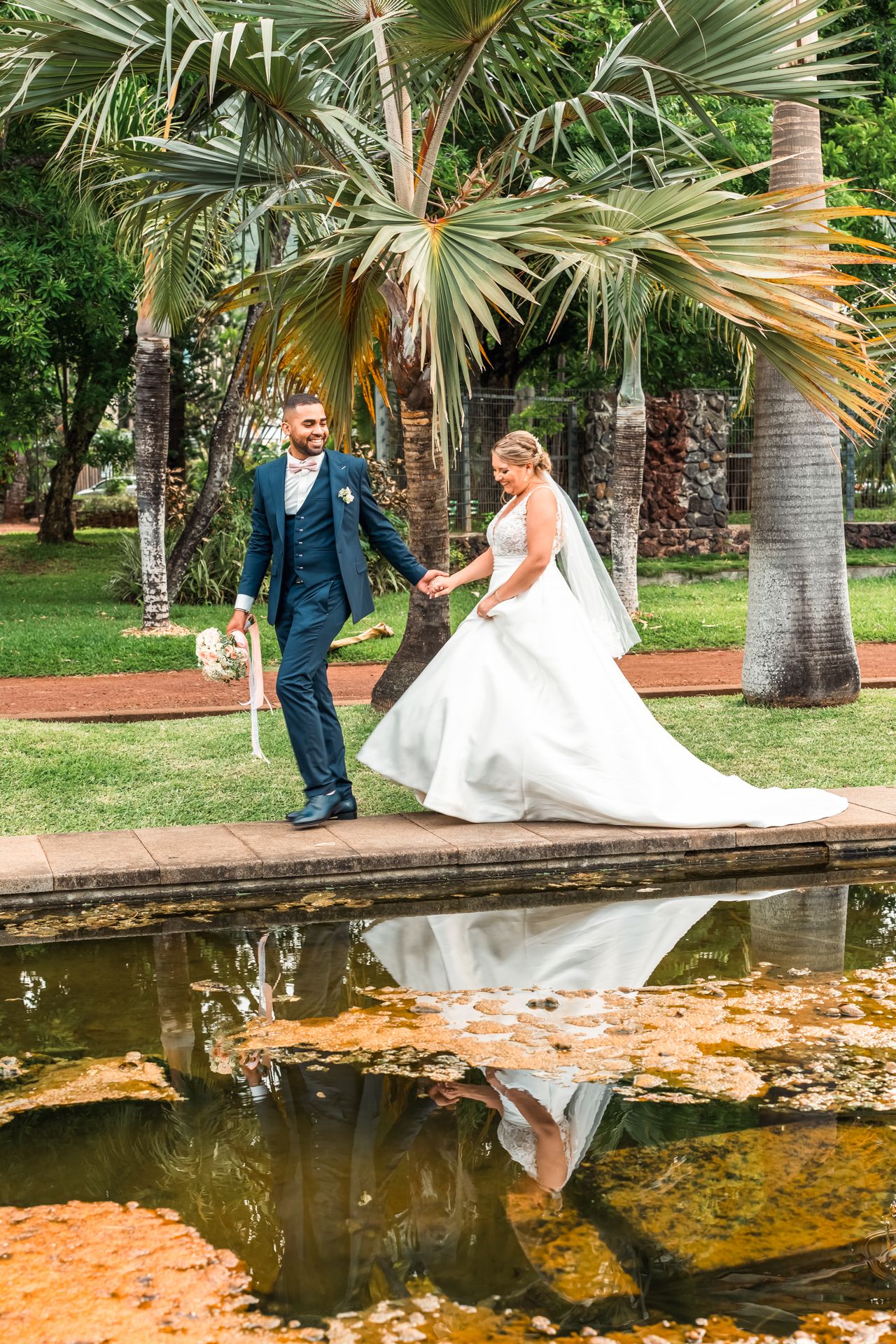 A newlywed couple strolling along a reflective pond surrounded by tropical palm trees in a lush garden setting.