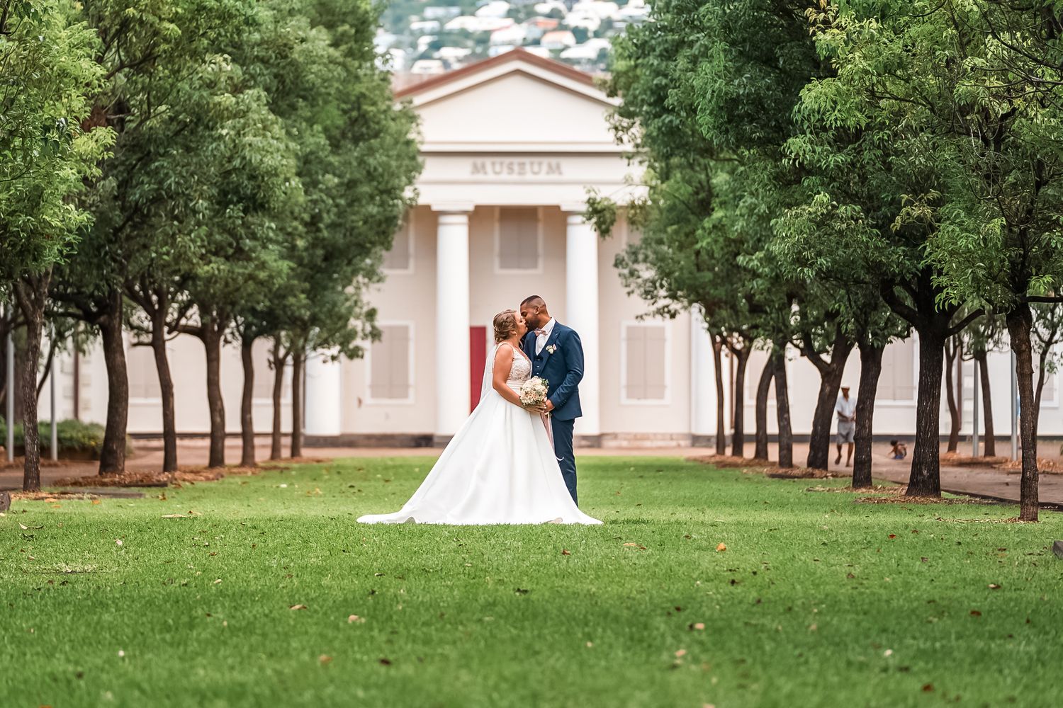 A couple shares a romantic moment in front of a neoclassical building surrounded by trees on a manicured lawn.