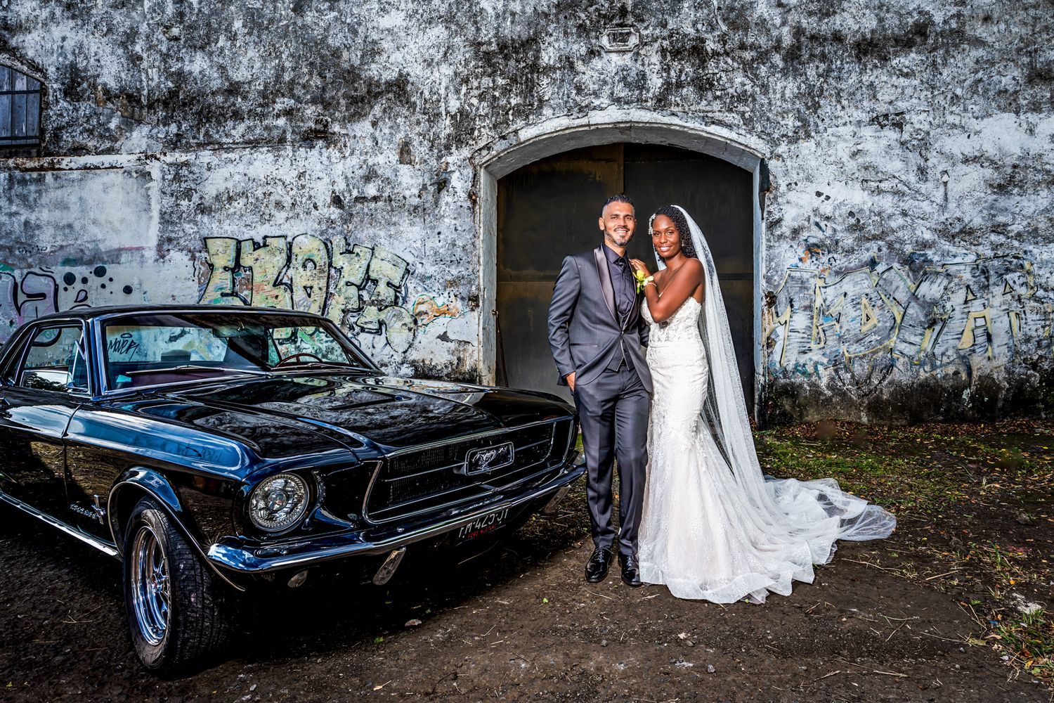 A classic black Mustang car parked next to a rustic stone archway during a wedding photo session.