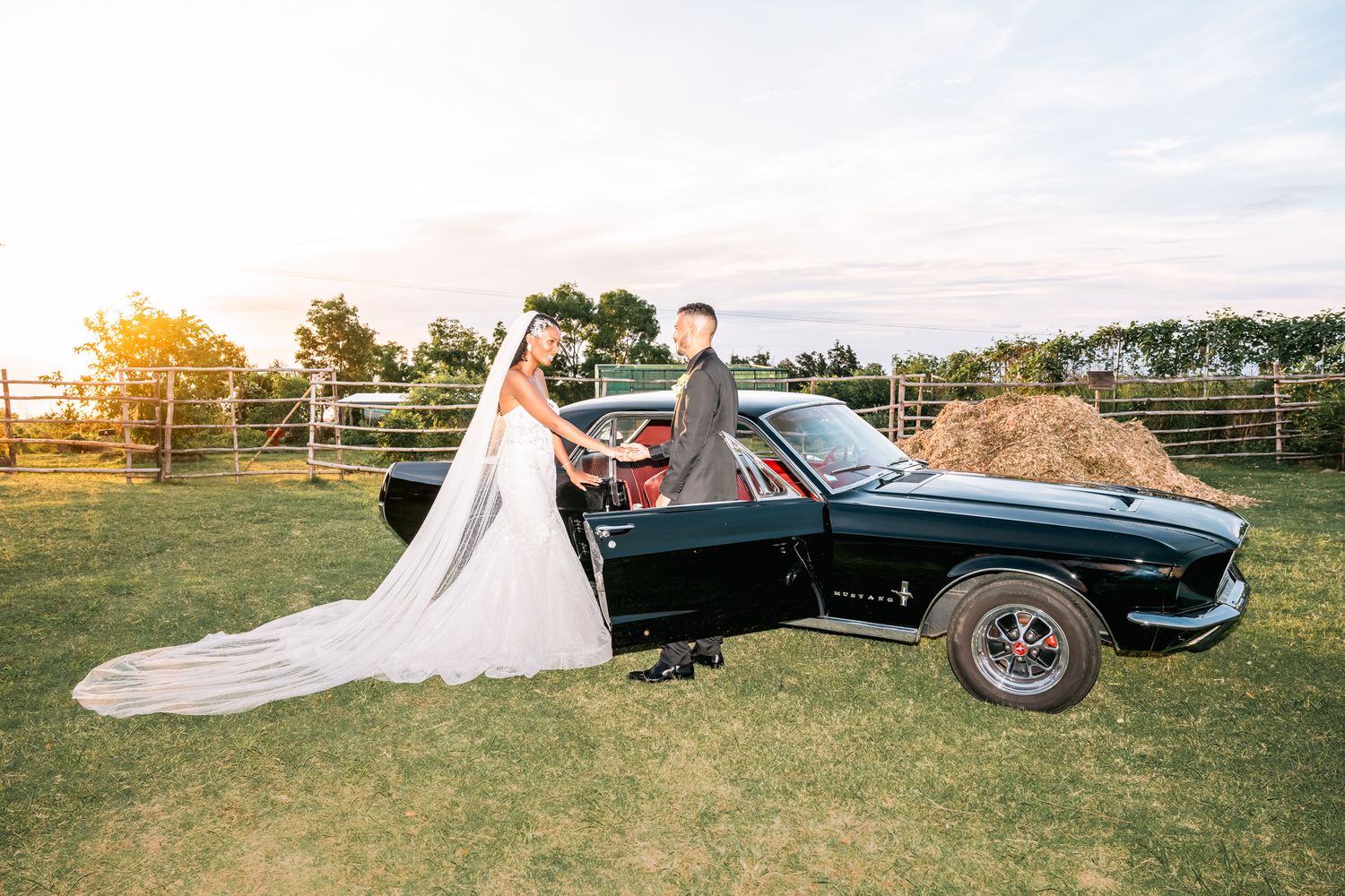 A classic black muscle car sits in a grassy field at sunset as a couple in wedding attire poses beside it.
