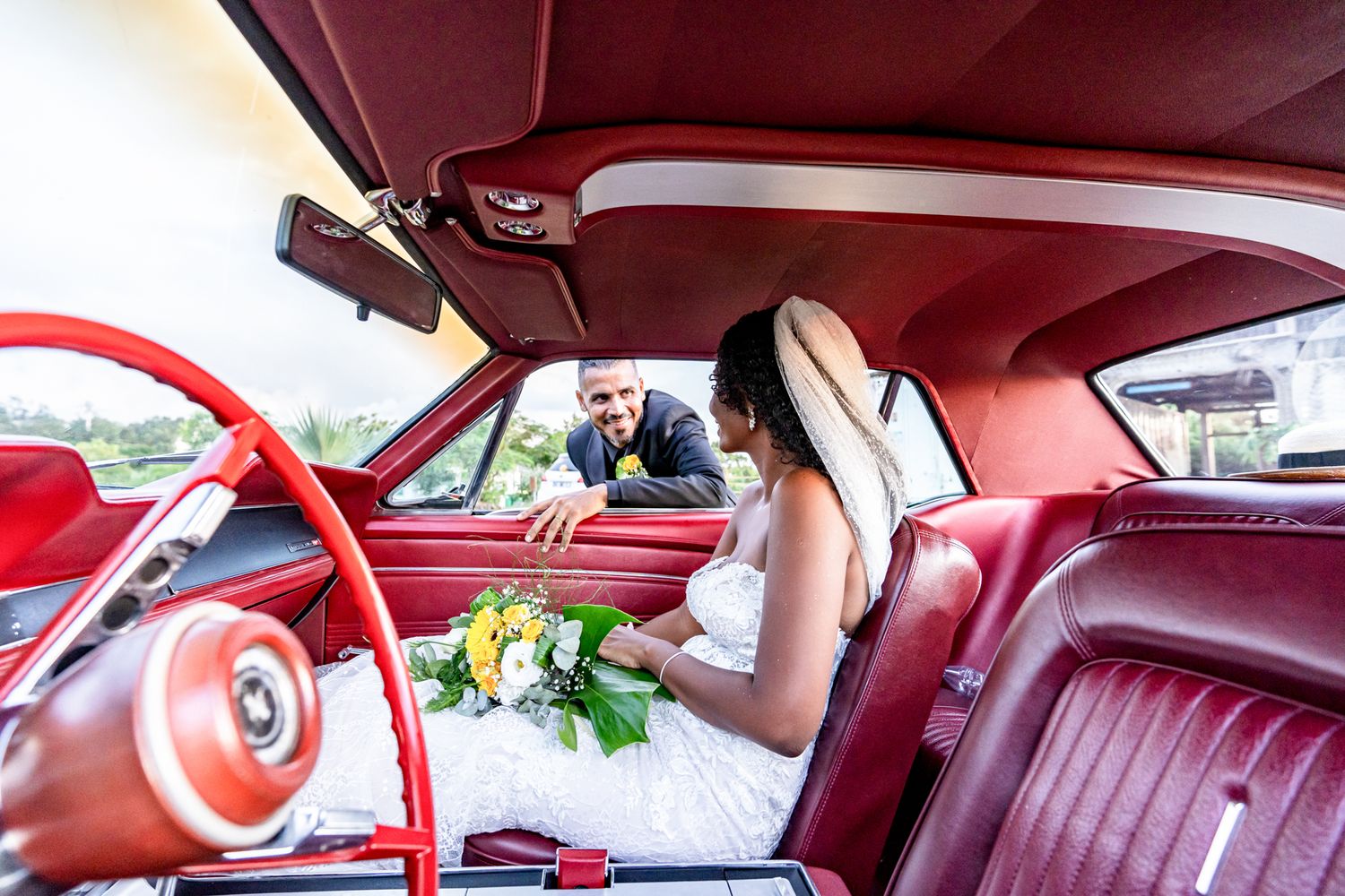 A classic car interior with red leather seats and steering wheel showing a bride with flowers in a vintage automobile.
