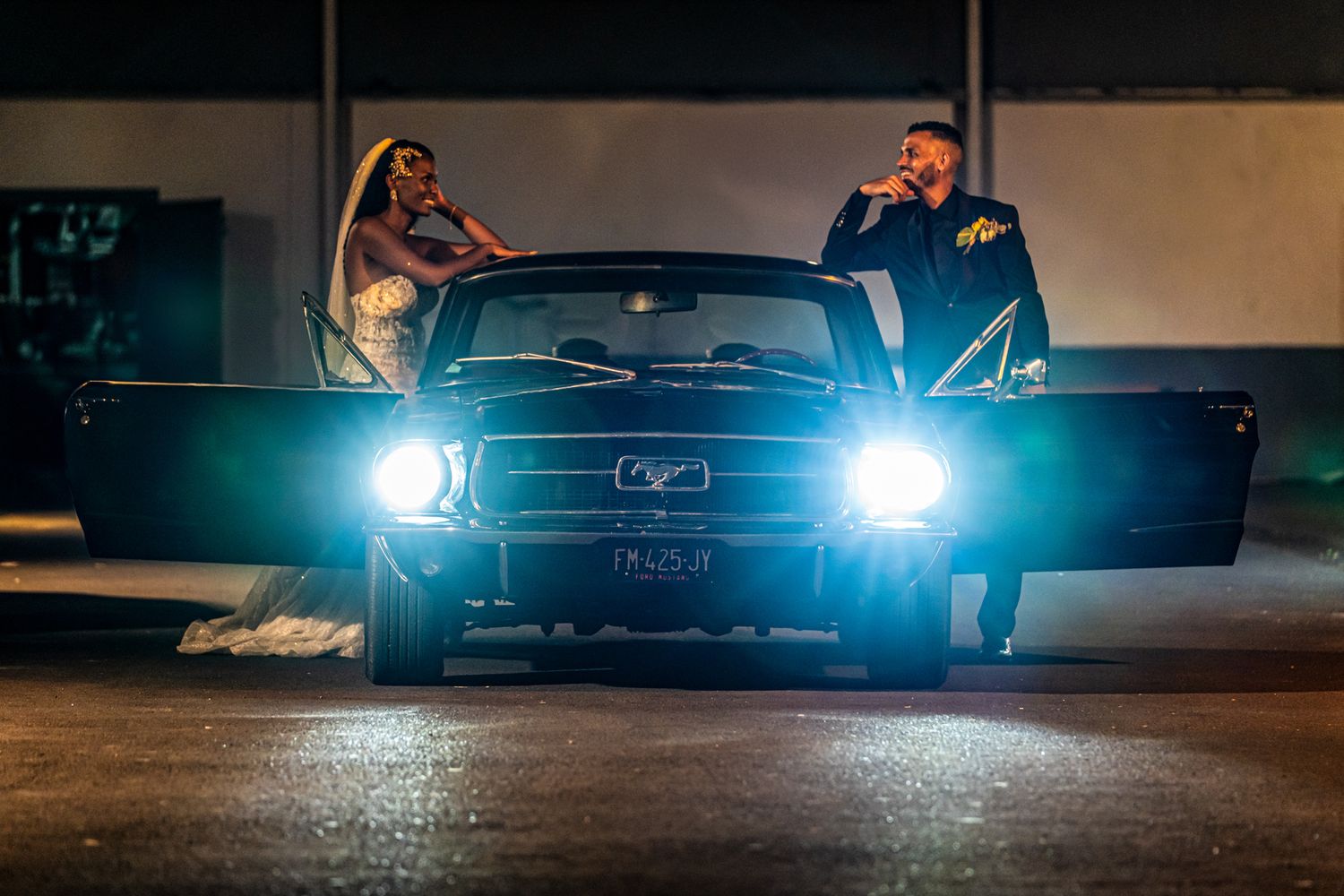 Two people pose with bright headlights shining from a classic Ford Mustang at night in a dark parking area.