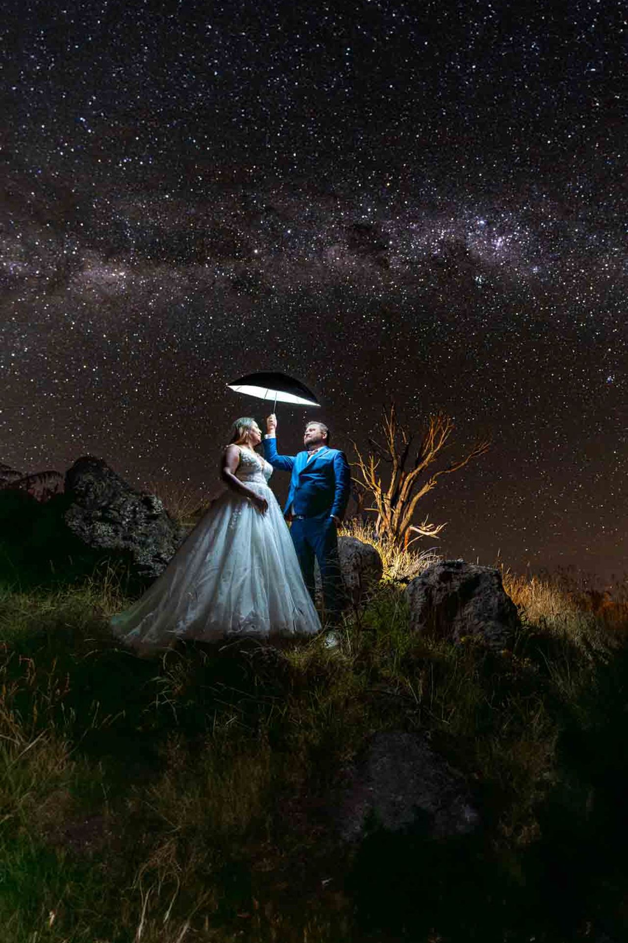 A couple in formal attire stands under an umbrella against a stunning starlit sky with the Milky Way visible above.