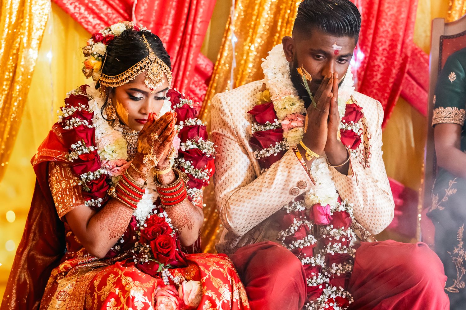 Traditional Indian wedding ceremony with couple wearing red and white attire adorned with flower garlands during prayer.