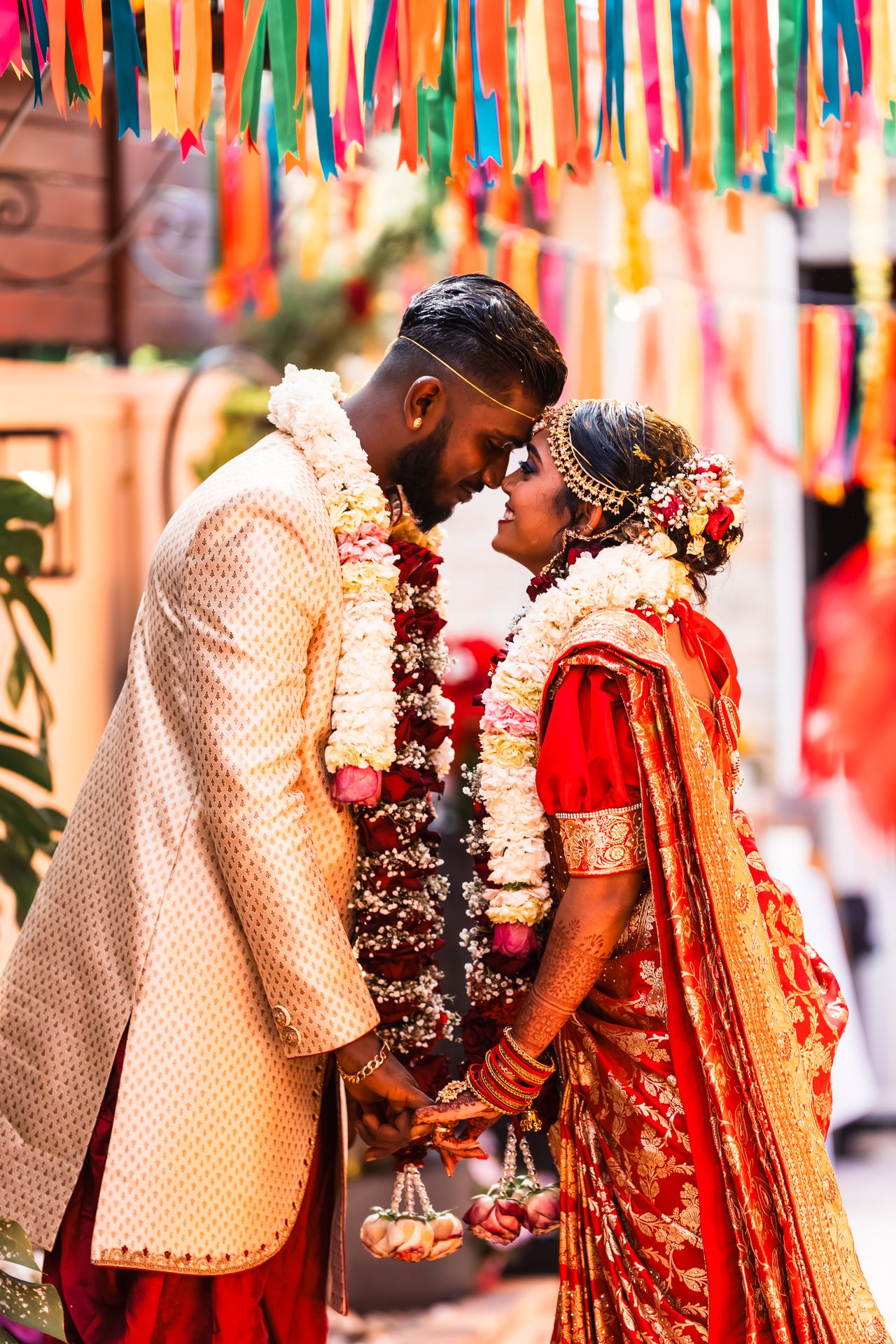 A joyous wedding couple in traditional Indian attire share a moment under colorful streamers at their celebration.