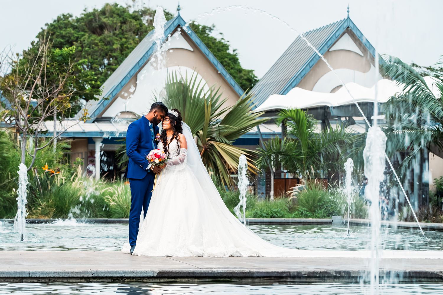 Wedding couple shares intimate moment by fountain with tropical white peaked pavilions in background.