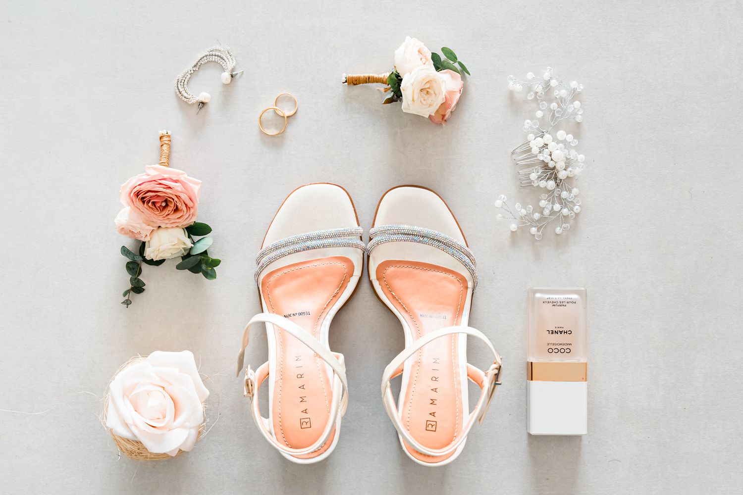 Flat lay of bridal accessories including peach sandals, roses, pearls, earrings and perfume bottle.