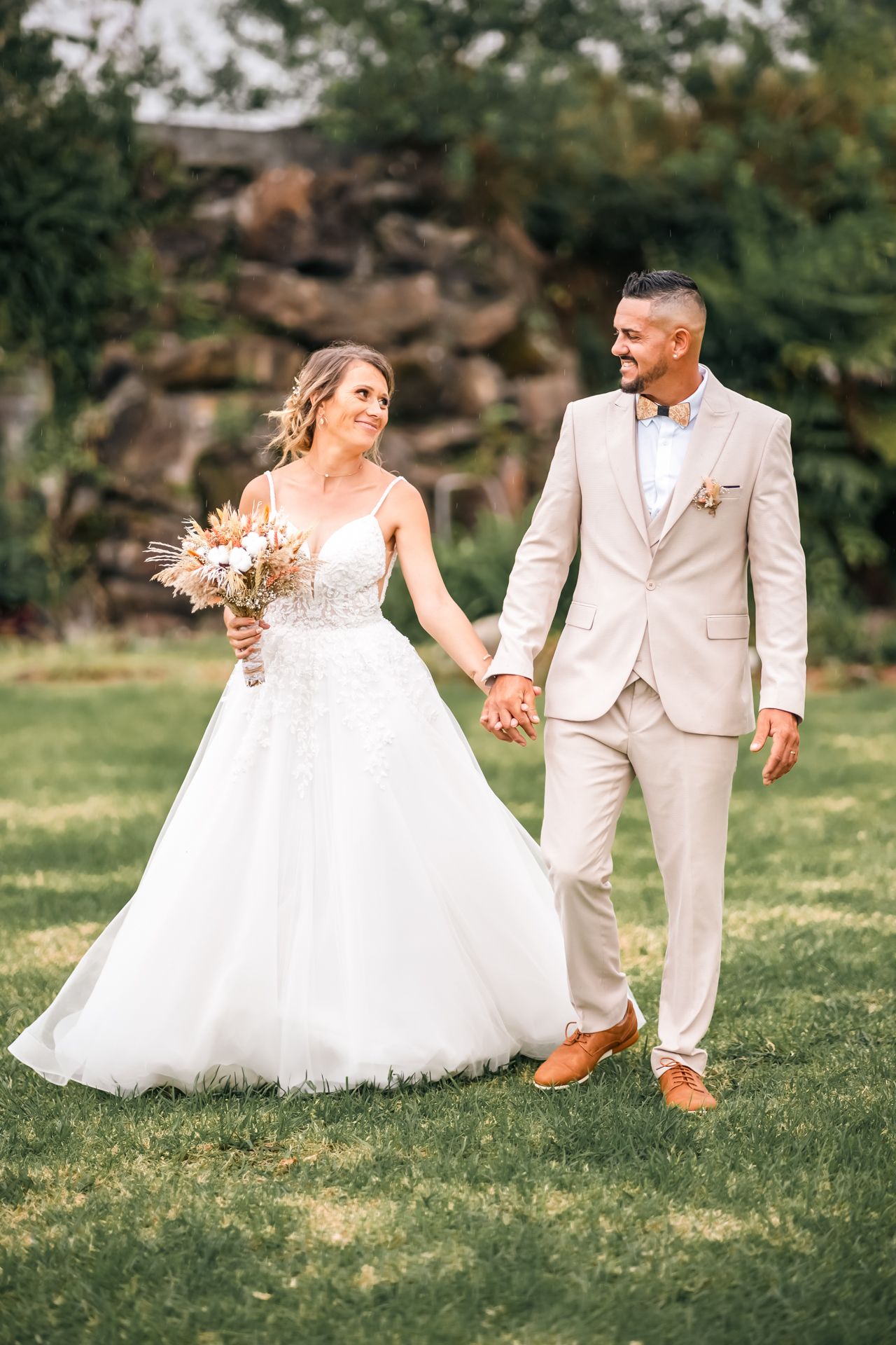 Newlyweds share a joyful moment on green lawn with scenic rock formations, bride in white gown and groom in beige suit.
