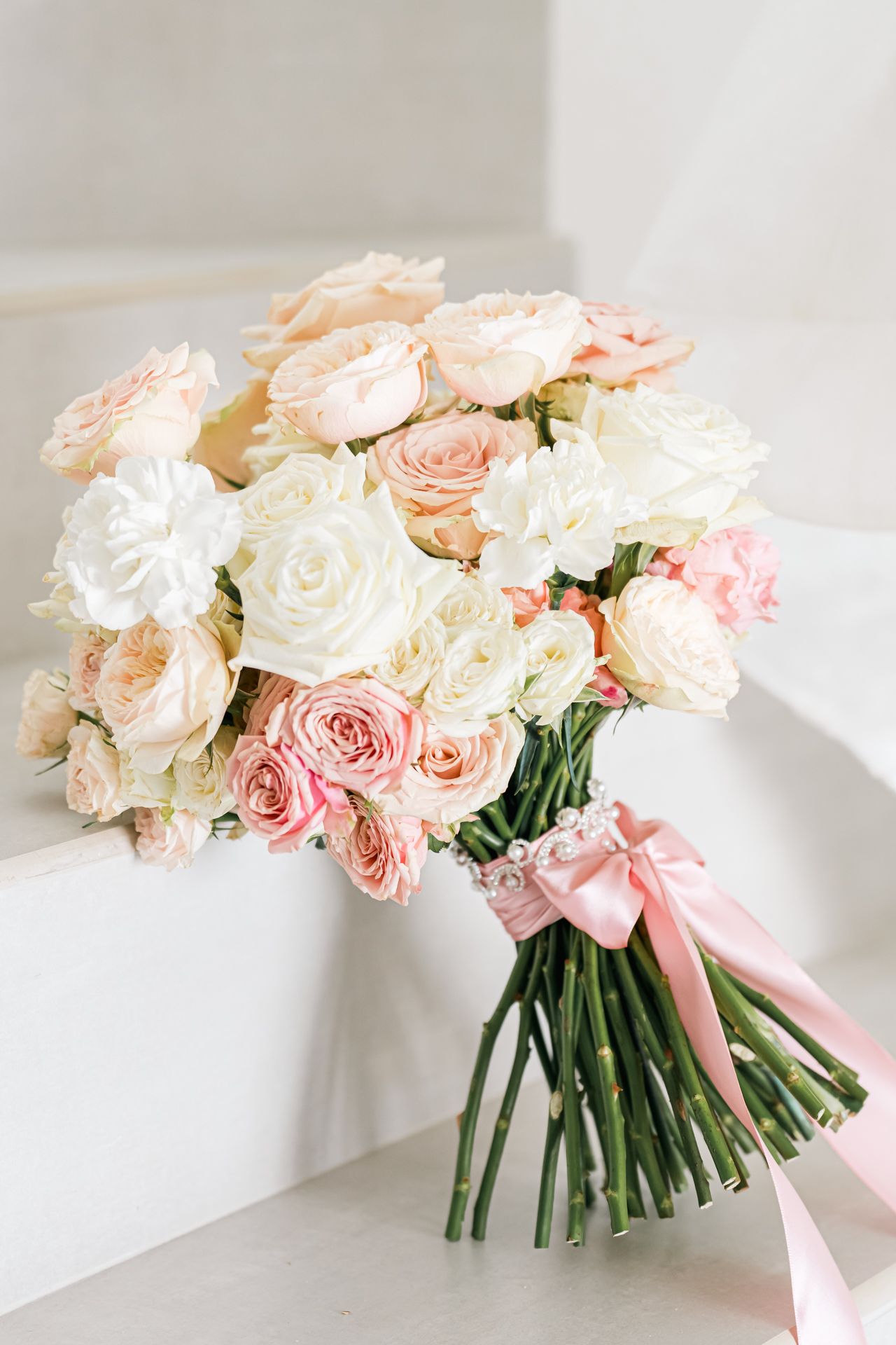 Elegant bridal bouquet of blush pink and white roses tied with a soft pink ribbon against a light background.