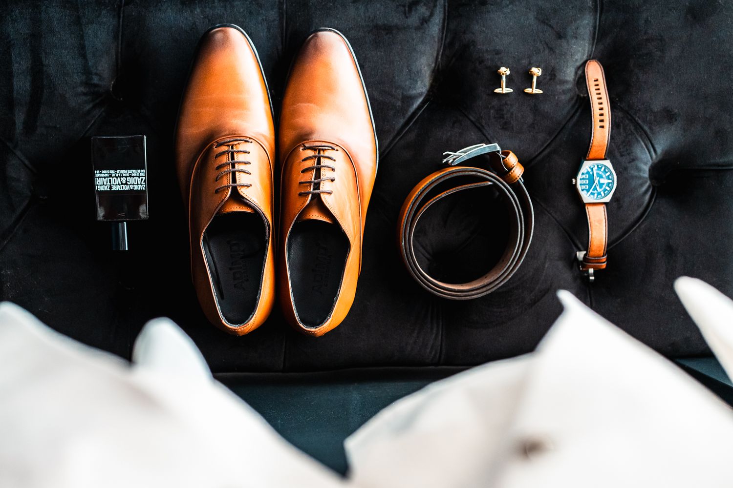 A flat lay of brown leather dress shoes, belt, watch, and cufflinks arranged on dark fabric.