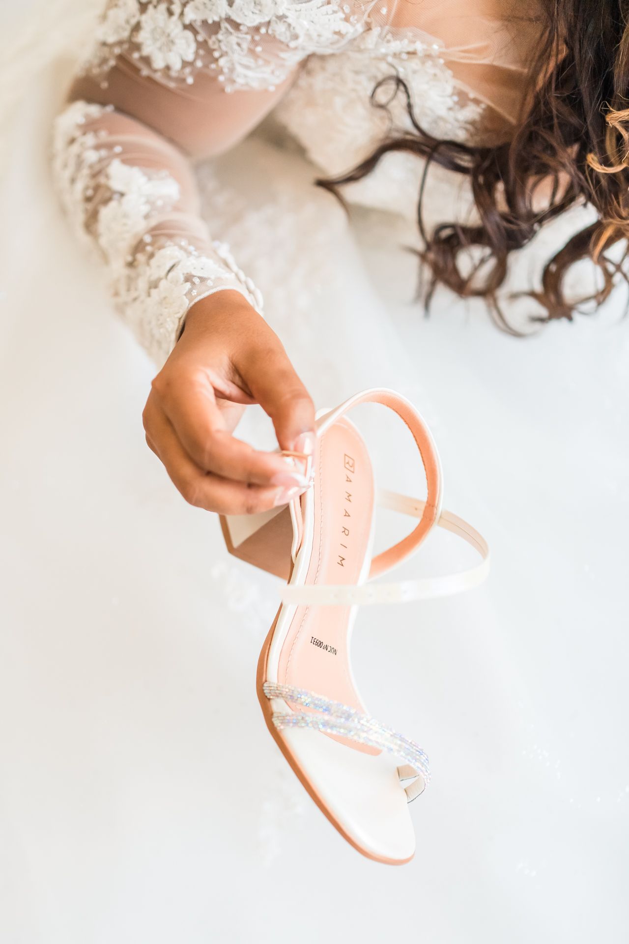 A hand in white lace bridal sleeves holds a delicate pink and white wedding sandal against a bright background.