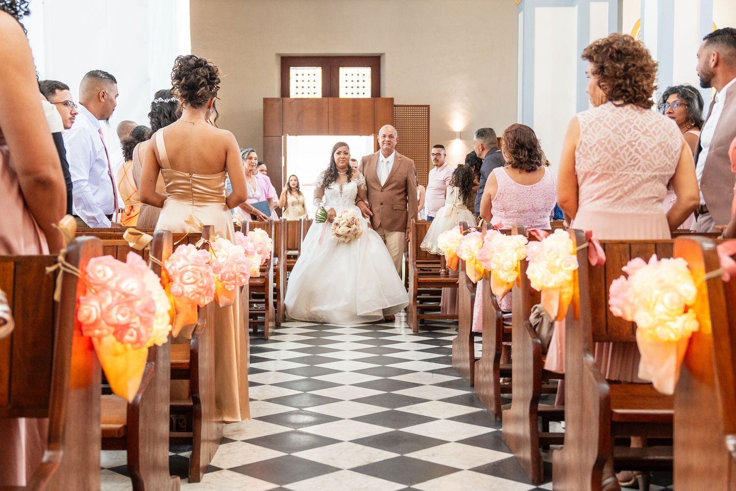 Wedding ceremony in a chapel with checkerboard floors and bridesmaids carrying peach-colored lantern bouquets down the aisle.