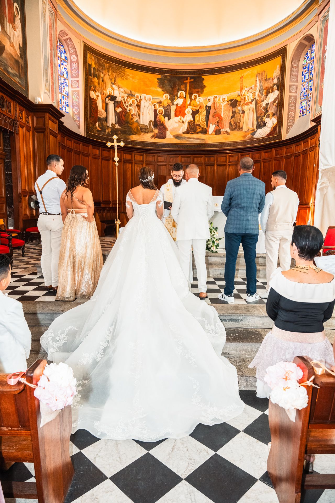 A bride in a flowing white gown with a long train stands at the altar during a wedding ceremony in a grand church.