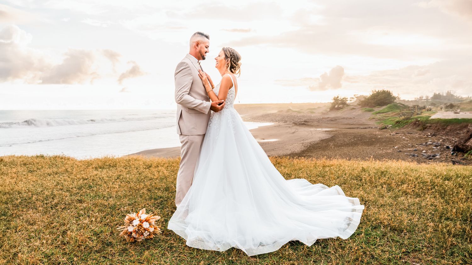 Wedding couple embraces on coastal cliff at sunset, with bride in flowing white dress and groom in light gray suit.