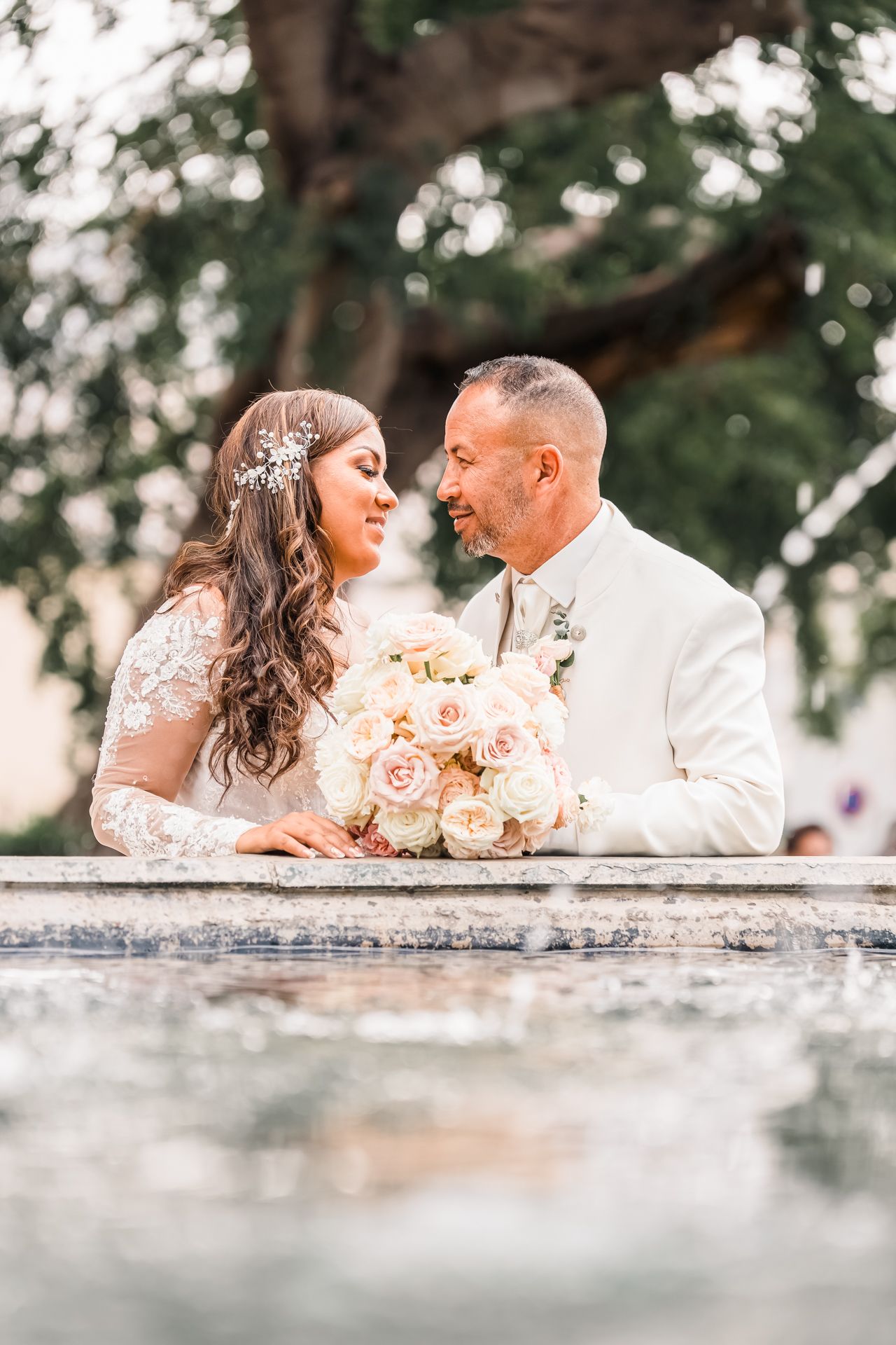 Couple in elegant white wedding attire share an intimate moment with peach rose bouquet in outdoor garden setting.