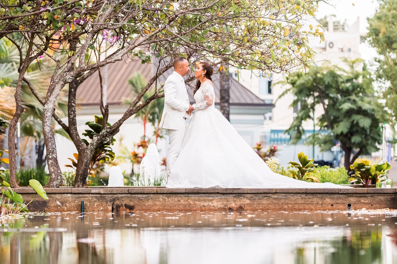 A romantic wedding scene beside a pond with bride and groom embracing on a stone wall surrounded by lush gardens.