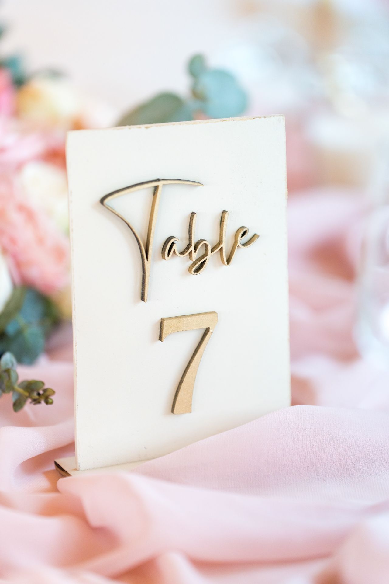 Elegant table number seven made of gold script lettering on white acrylic with pink fabric backdrop and floral accents.