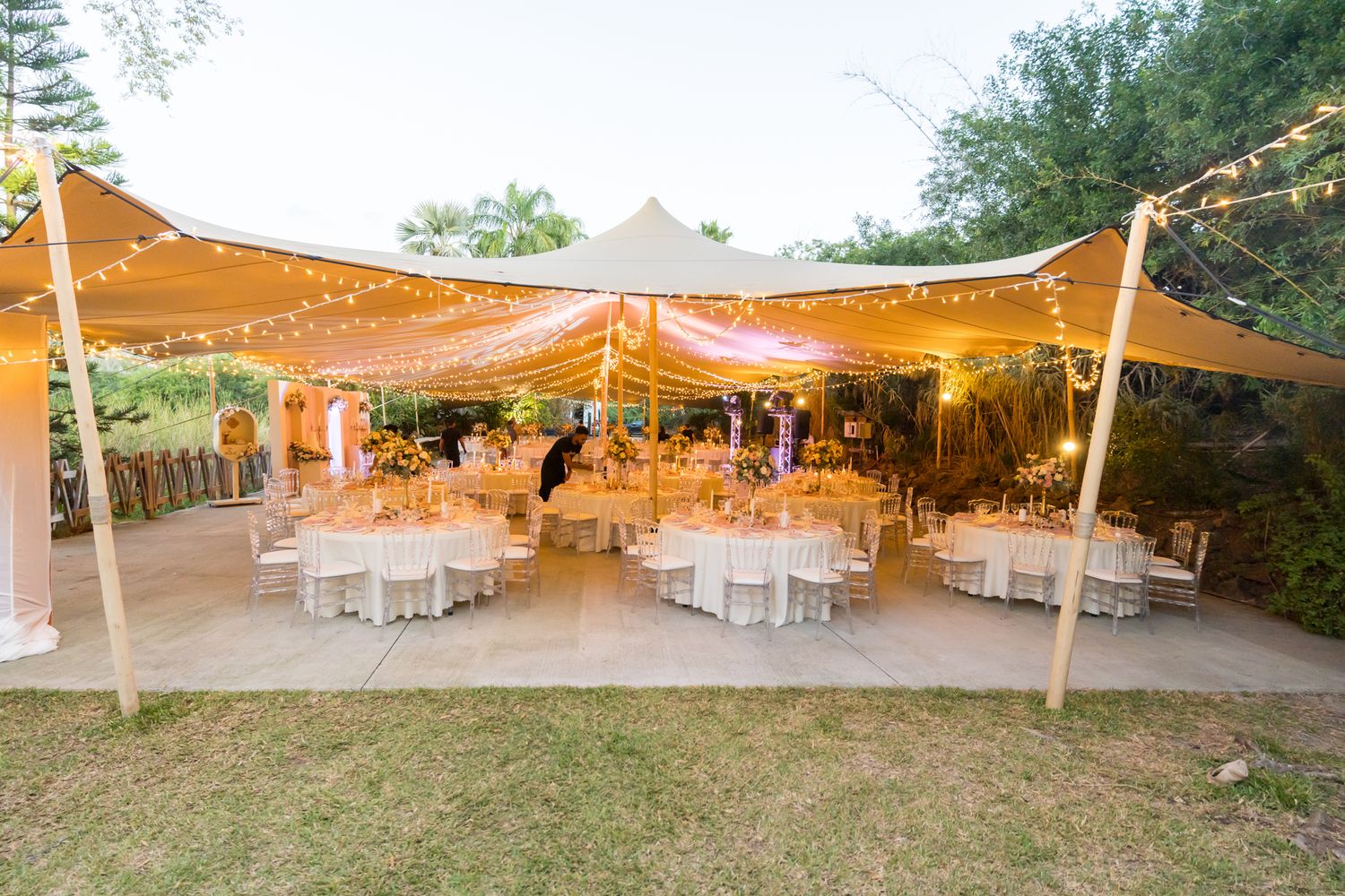 Outdoor wedding reception setup with white tables under a large tan canopy decorated with string lights at dusk.