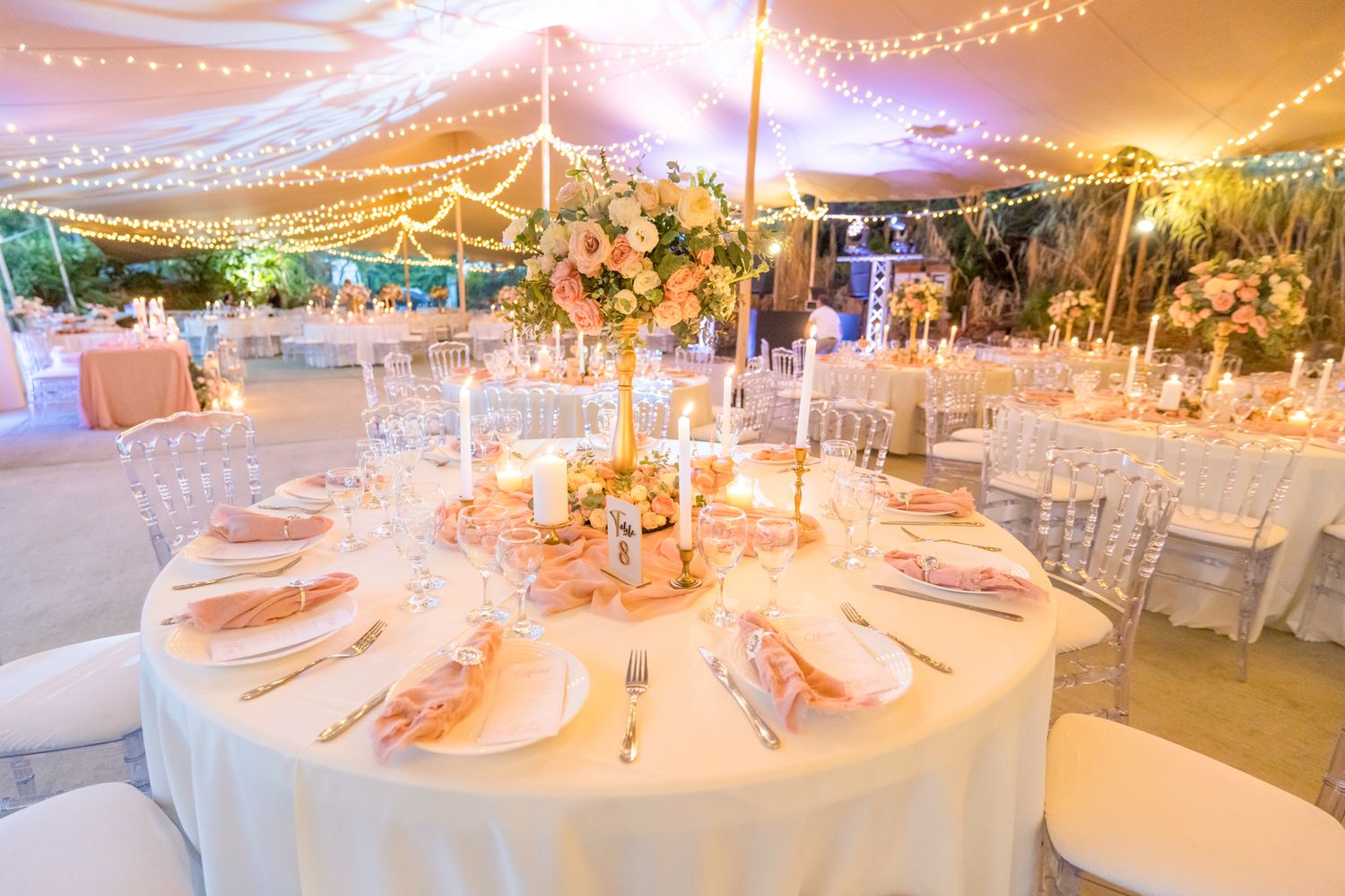Elegant wedding reception table setup with white linens, string lights overhead and floral centerpiece at dusk.